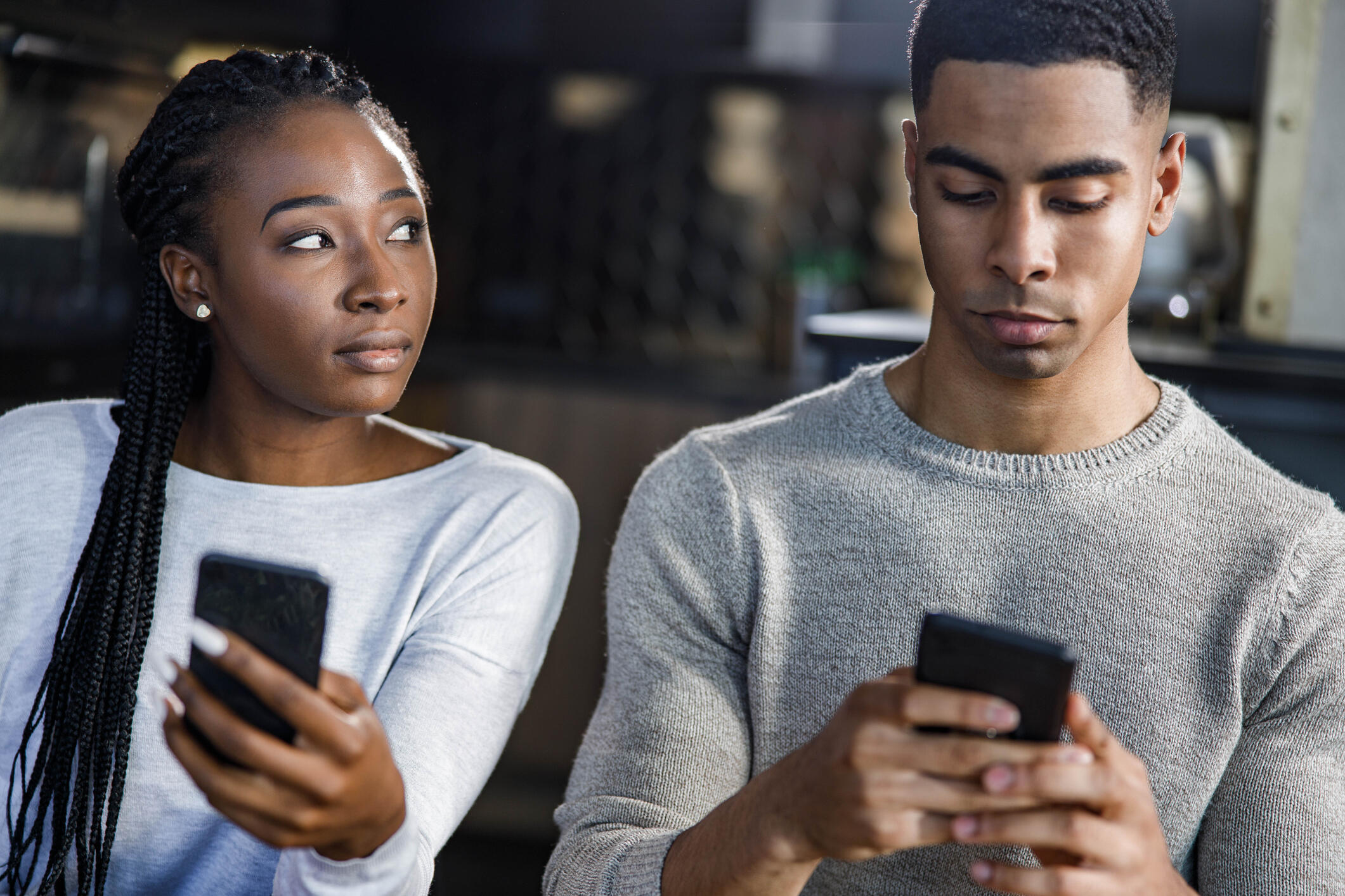 A photo of a man and woman sitting next to each other. Both people are holding cell phones, but the woman is looking at the man to her right supsicously while the man is looking at his phone. 
