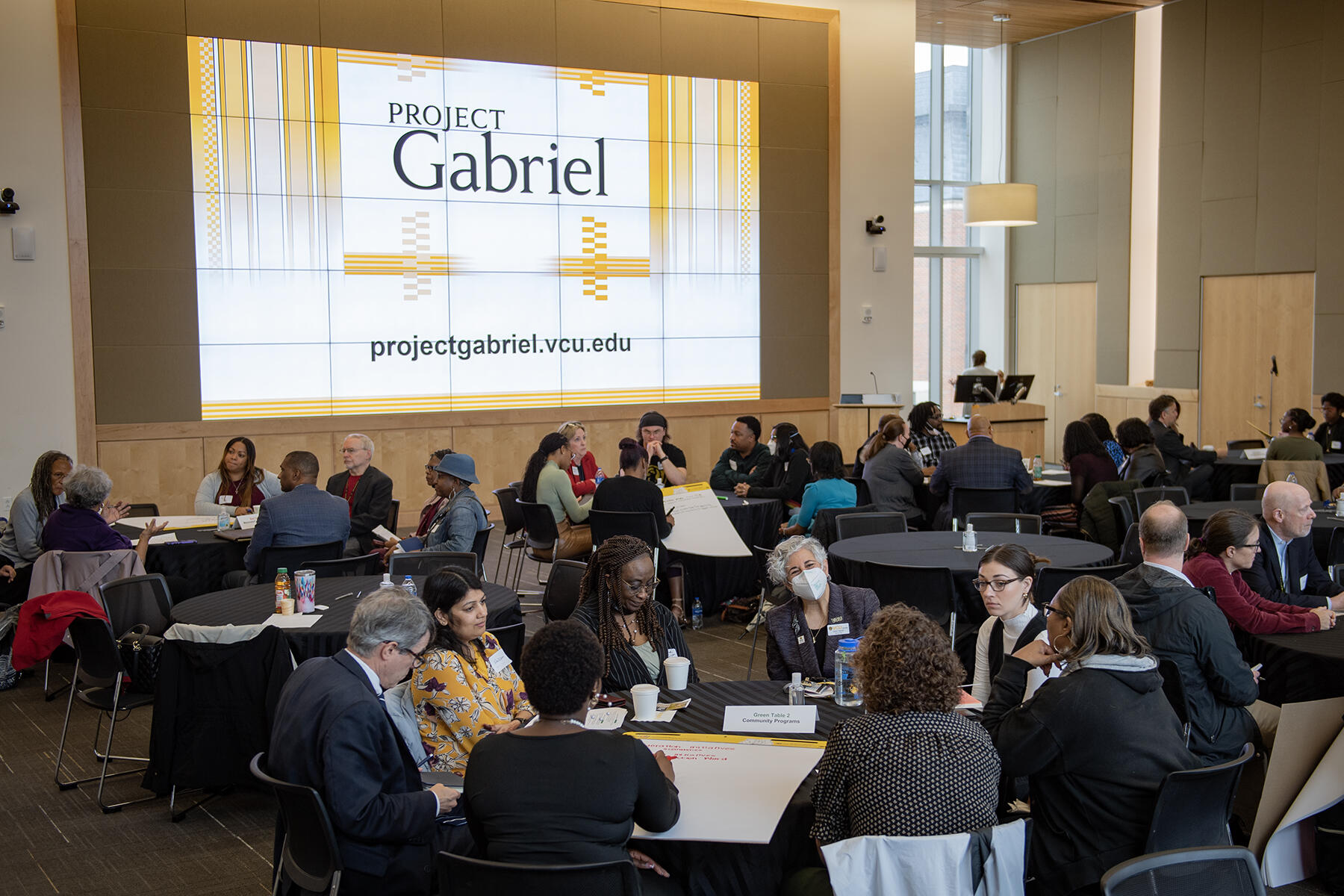 People sitting at circular tables in a large room. There is a screen that says \"PROJECT Gabriel\" behind them. 