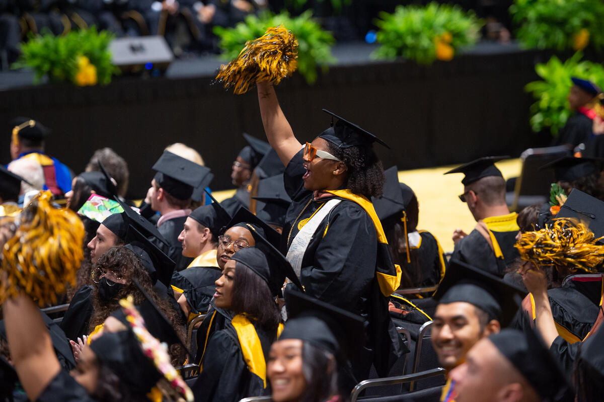 A group of people sitting in chairs wearing graduation caps and gowns. One woman stands in the middle of the crowd, cheering and waving a pompom in the air with her right arm. 