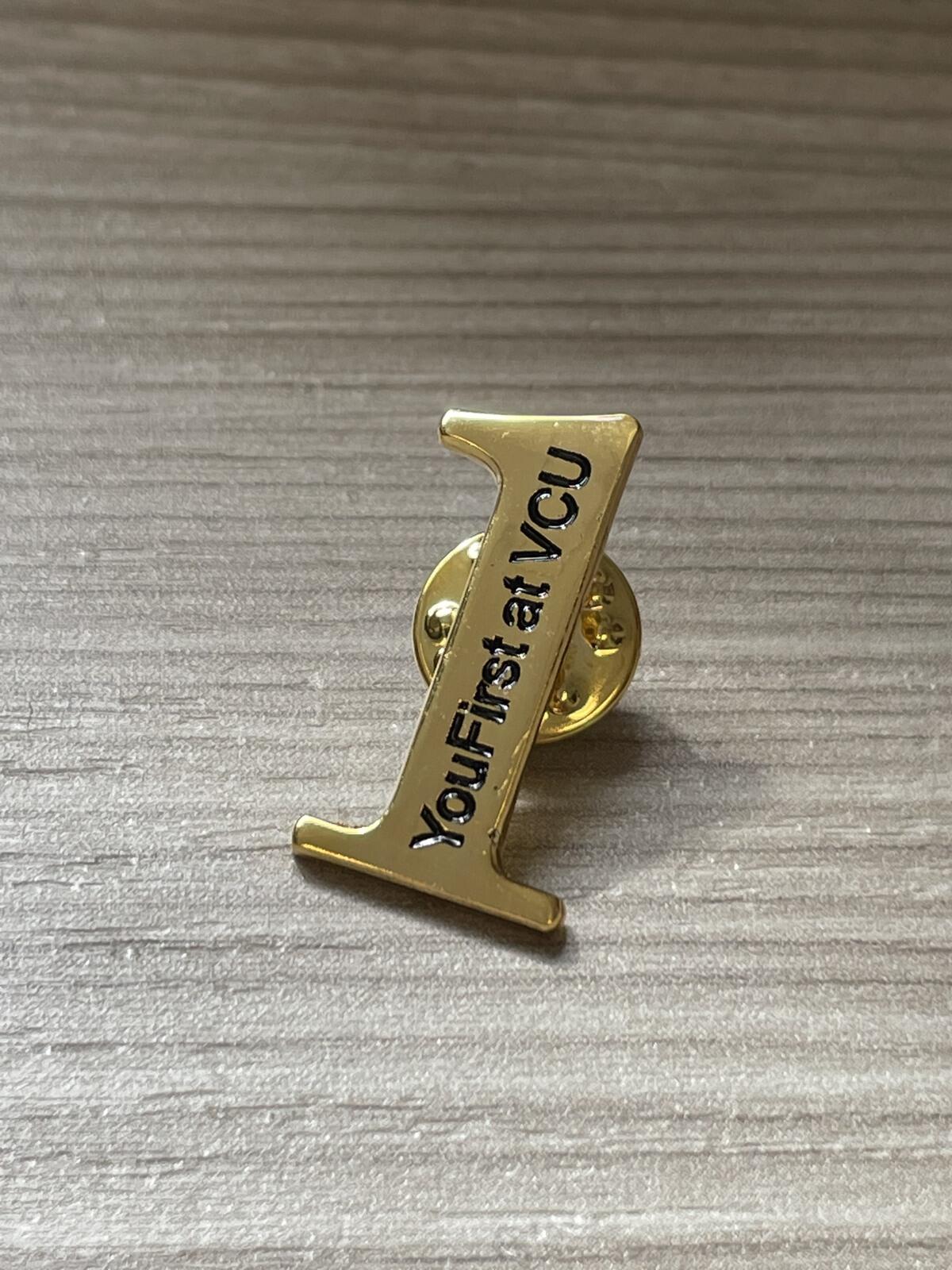 A photo of a gold pin shaped in the number one. Black text on top of the pin reads \"YouFirst at VCU\"