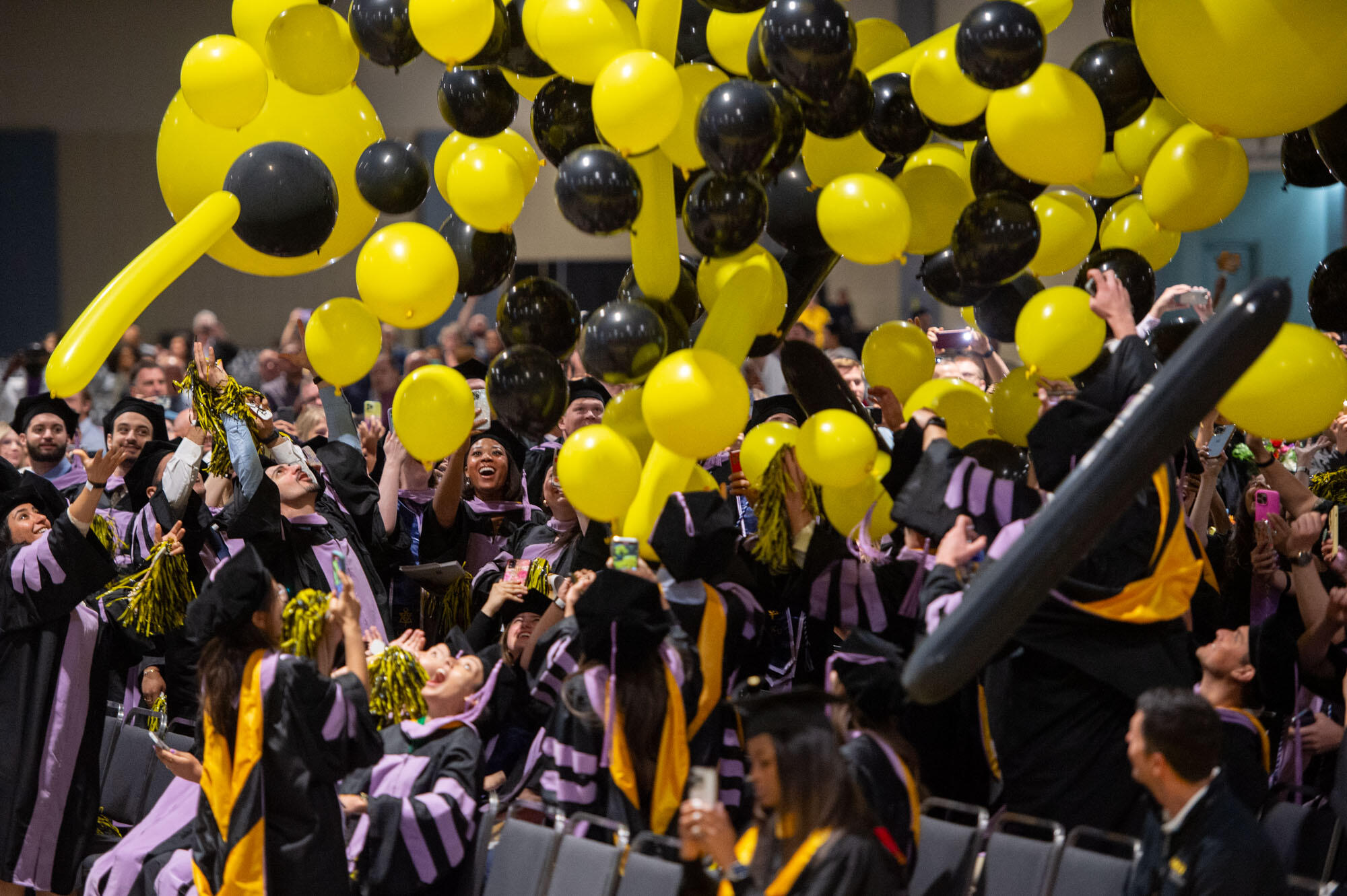 A photo of a crowd of students wearing graduation caps and gowns standing up under a bunch of falling balloons. The balloons are black and yellow.