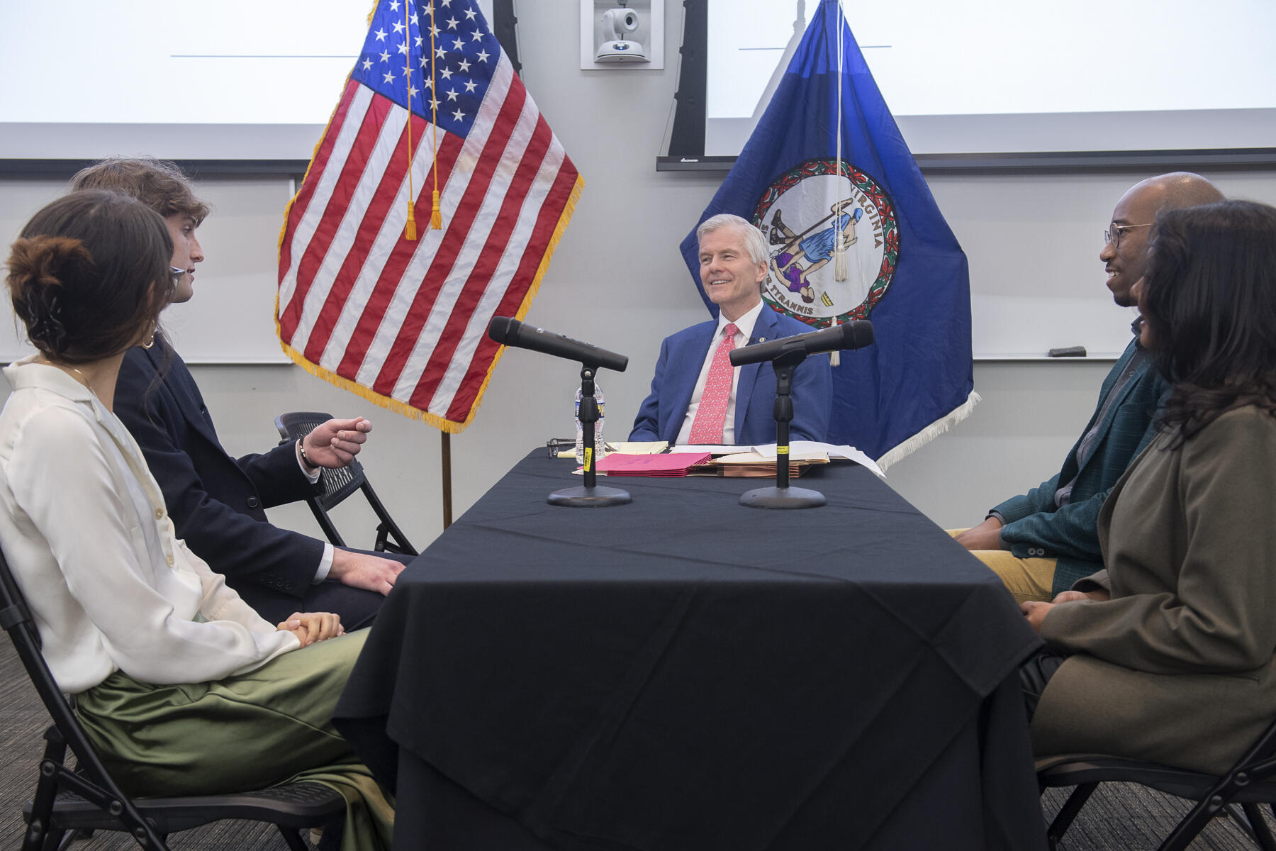 A photo of five people sitting around a rectangle shaped table. Two people sit on the left and right sides of the tame and a man sits at the end of the table. Behind the man is an American flag and a Virginia state flag. 