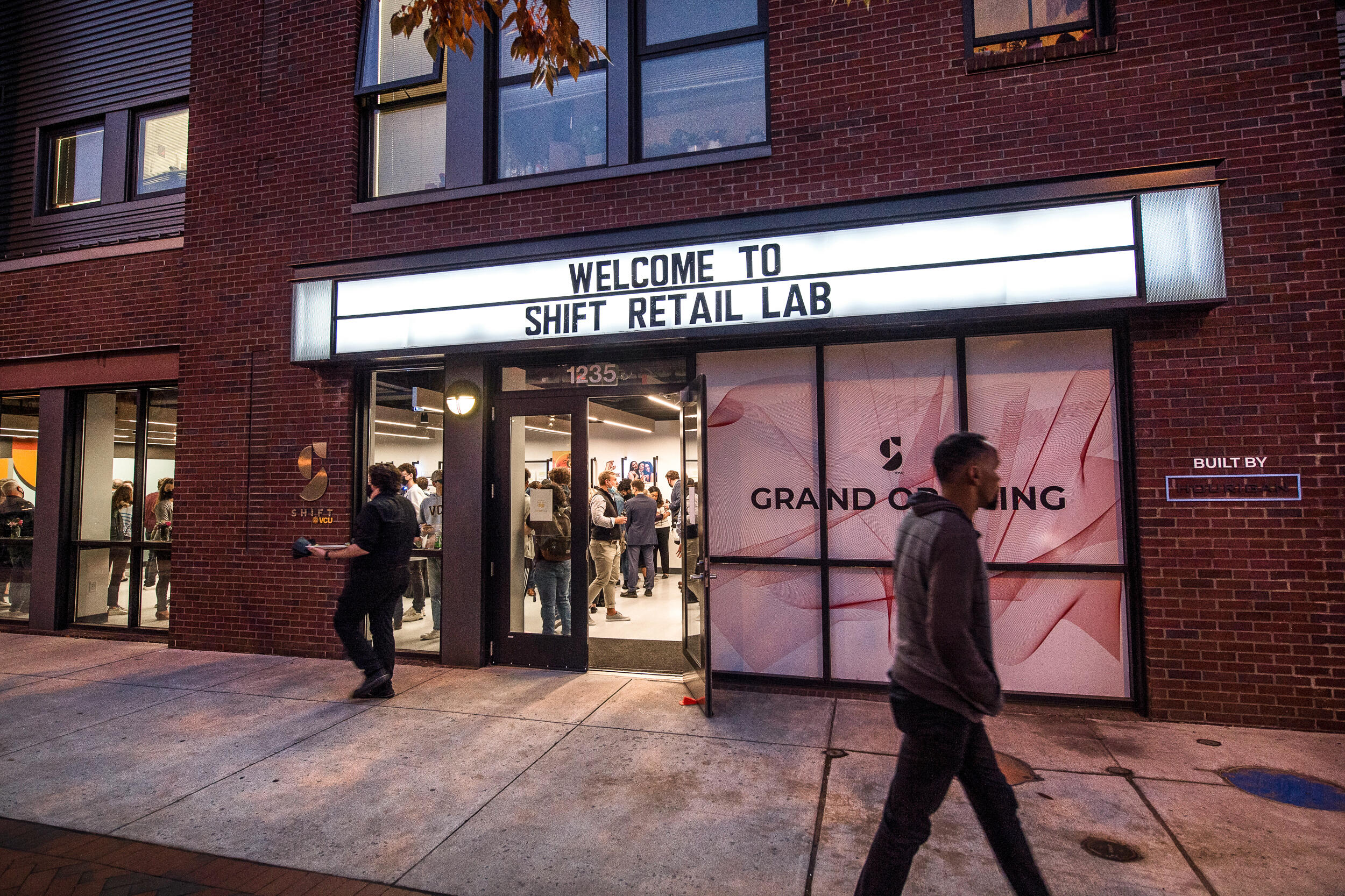 A person walks past the outside of a storefront on an urban street with a large brightly-lit marquee that says \"Welcome to Shift Retail Lab\"