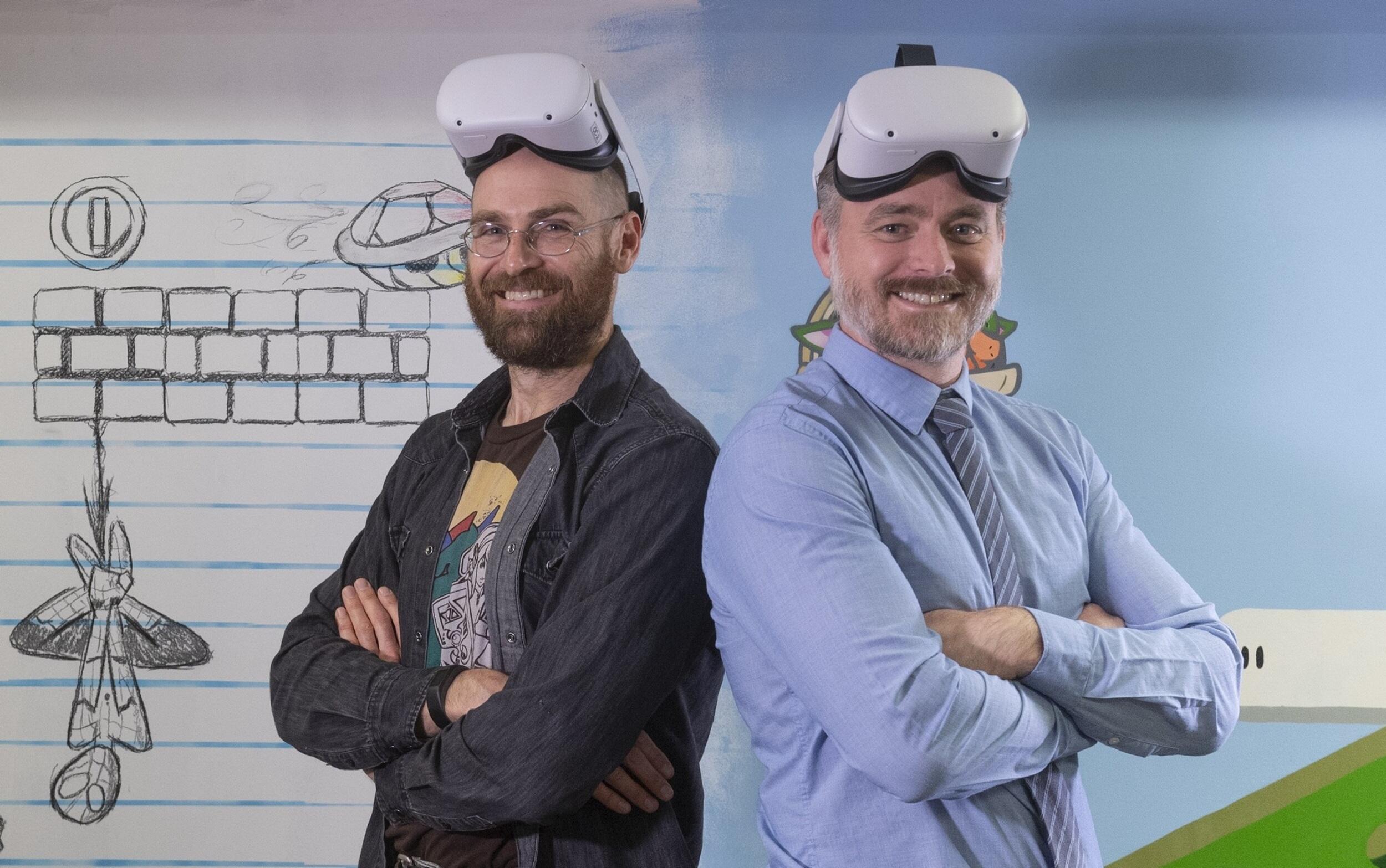 Two smiling men standing back to back with their arms crossed wearing virtual reality goggles headsets on their foreheads 