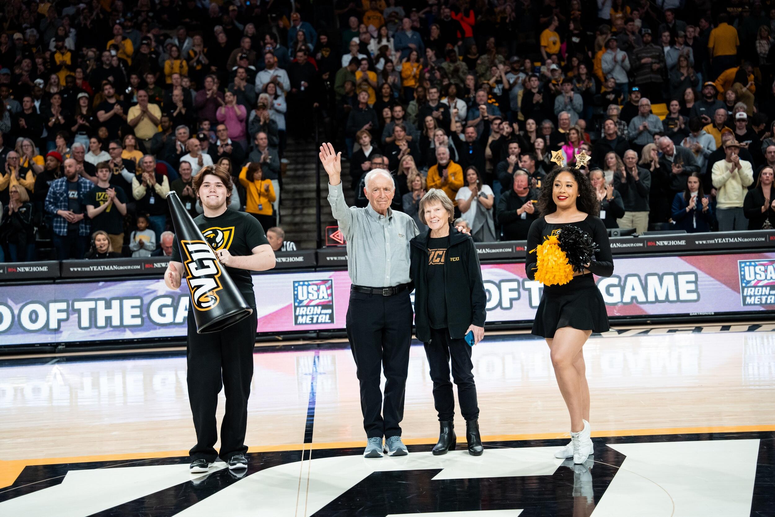 A photo of a man standing to the left of a woman with the arm closest to her behind her back and the other stretched up in the air waving. To the man's left is a another man holding a triangle flag that says \"VCU\" and to the woman's right is a another woman holding pompoms. Behind them, the stadium's seats are filled with people watching. 