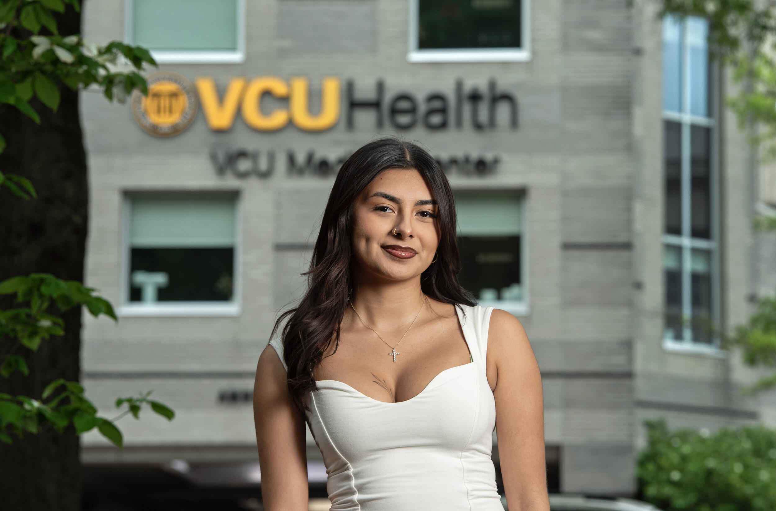 A photo of a woman from the waist up standing in front of a building that says \"VCU Health VCU Medical Center\" on it. 