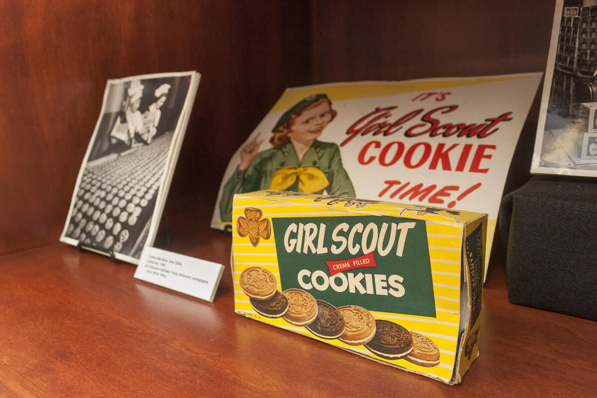 A box that has an illustration of cookies on it that says \"GIRL SCOUT COOKIES.\" Behind the box is an illustration of a girl scout with the text \"It's Girl Scout COOKIE TIME!\" written on it. Next to the box and illustration is a black and white photo of two workers in a factory with a cookies in front of them. 