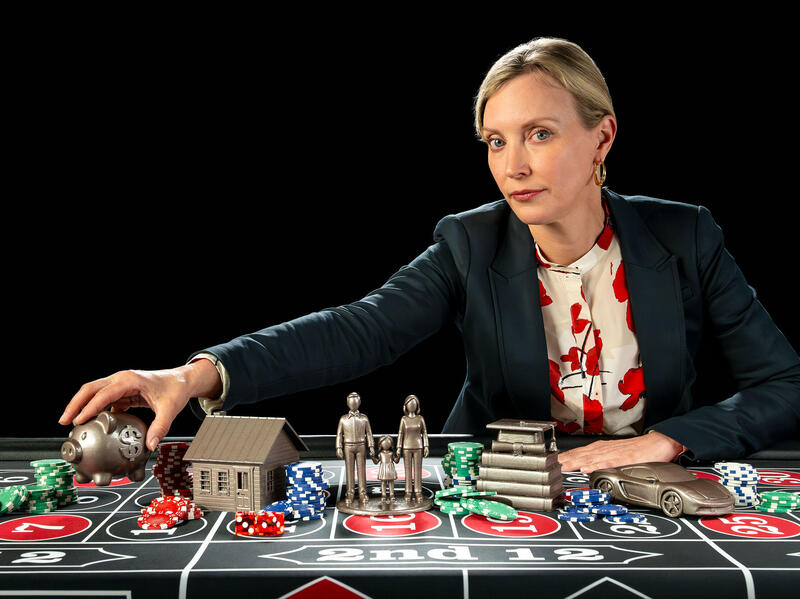 Carolyn Hawley reaches across a gambling table filled with poker chips and small statues of a house, people, a stack of books and a car; she holds a piggy bank. 