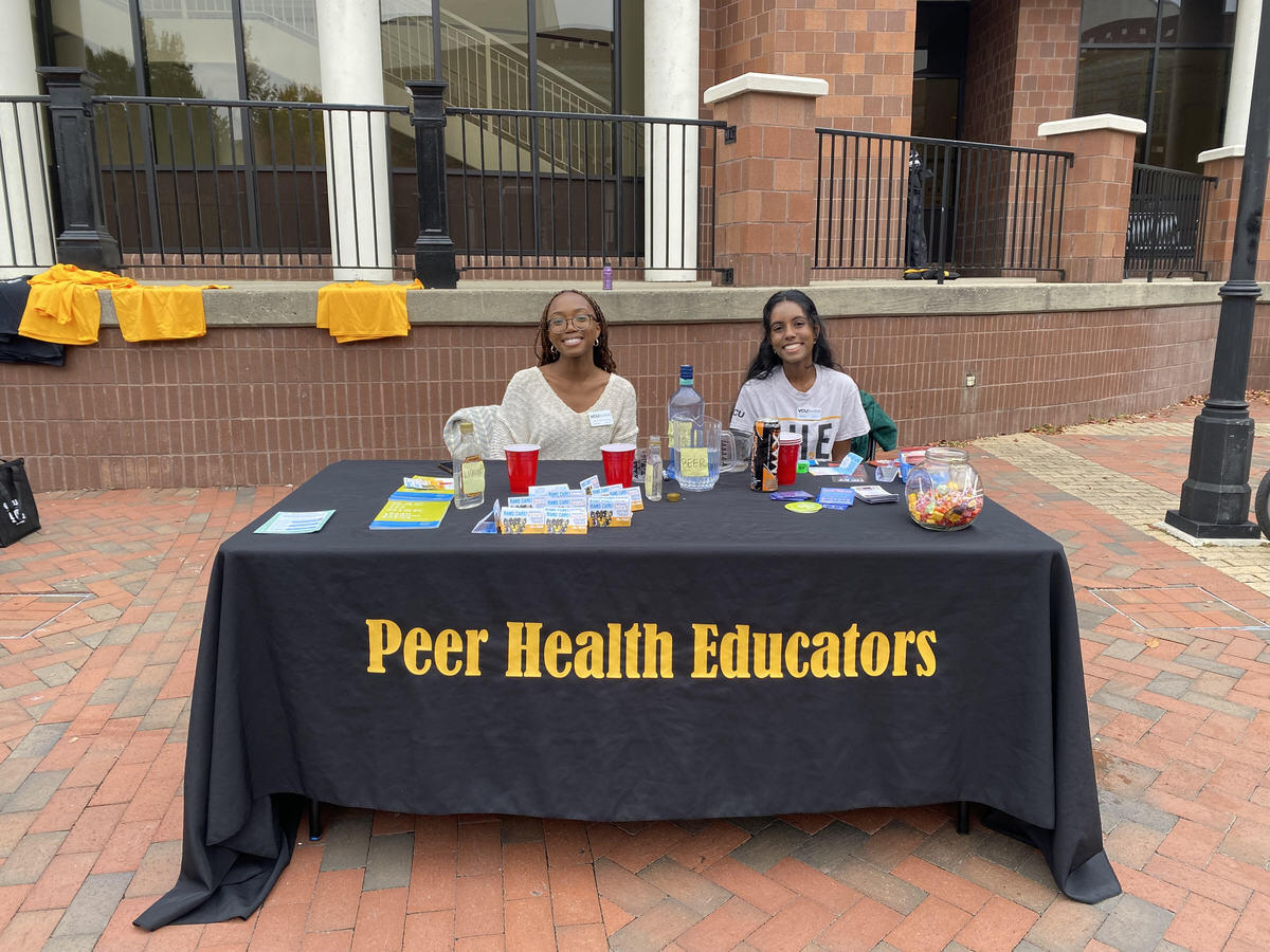 Two women smiling and sitting at a table with a table cloth that says \"Peer Health Educators\" across the front. 