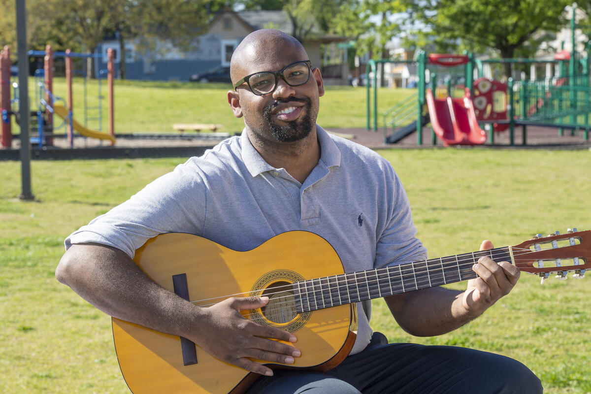A man sitting in the grass holding an acoustic guitar. Behind him is a playground. 