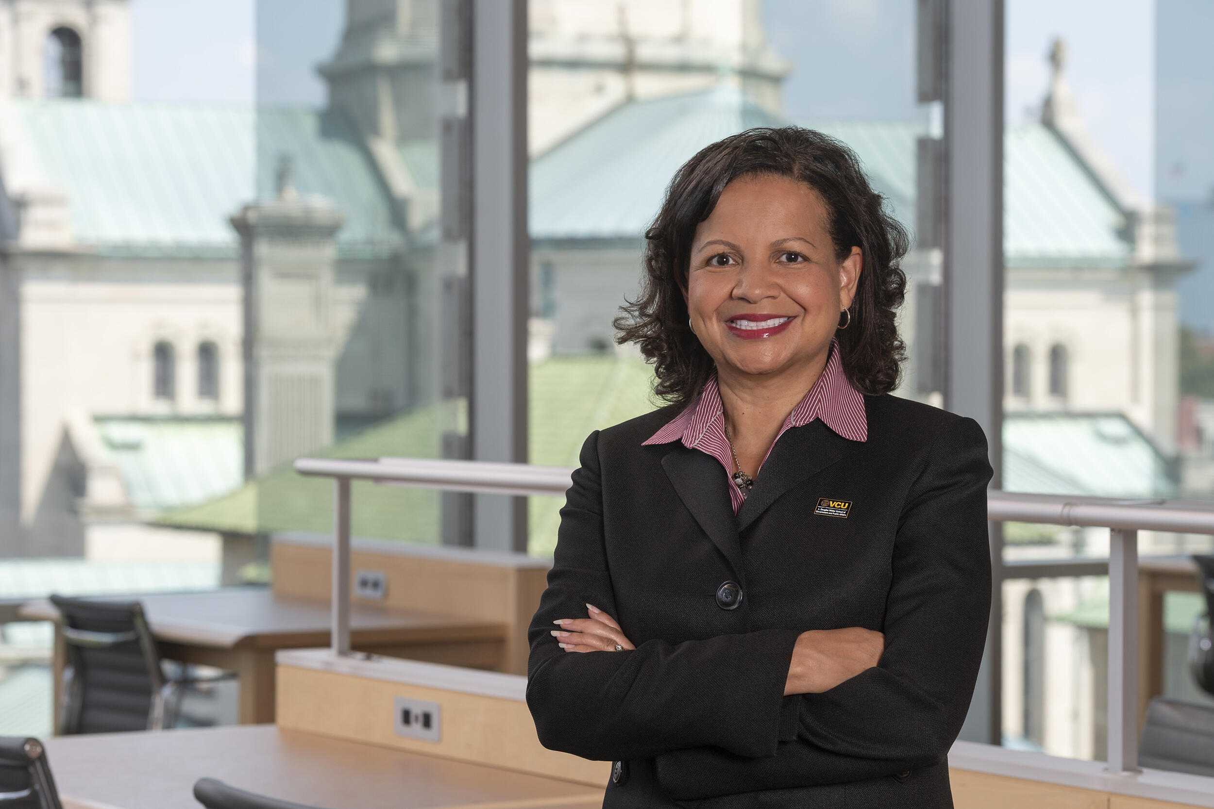 A photo of a woman from the waist up. She is standing in front of a window with her arms crossed against her chest. 