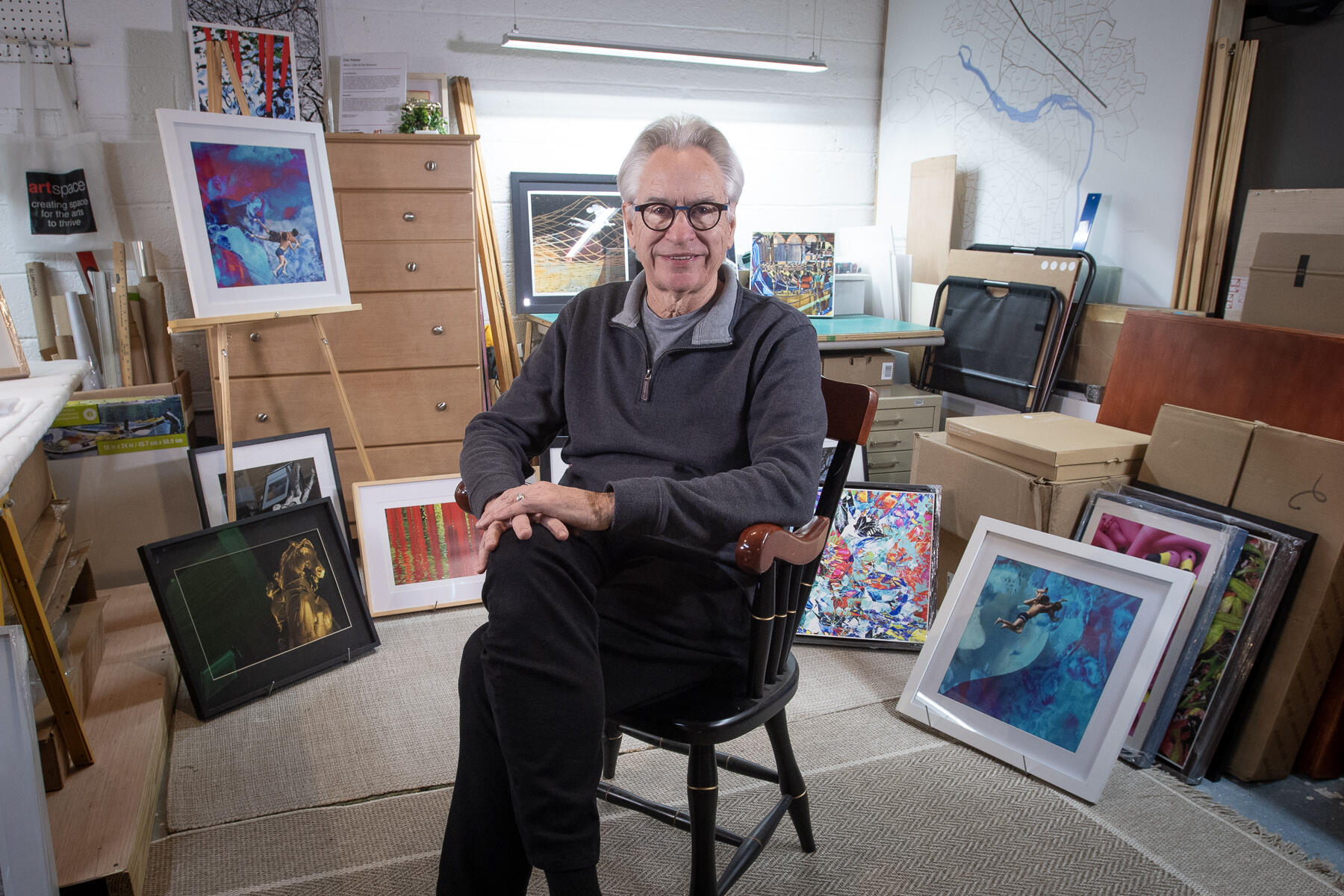 A photo of a man sitting in a chair in the middle of a room. Around him are several paintings sitting against boxex and on an easle. 