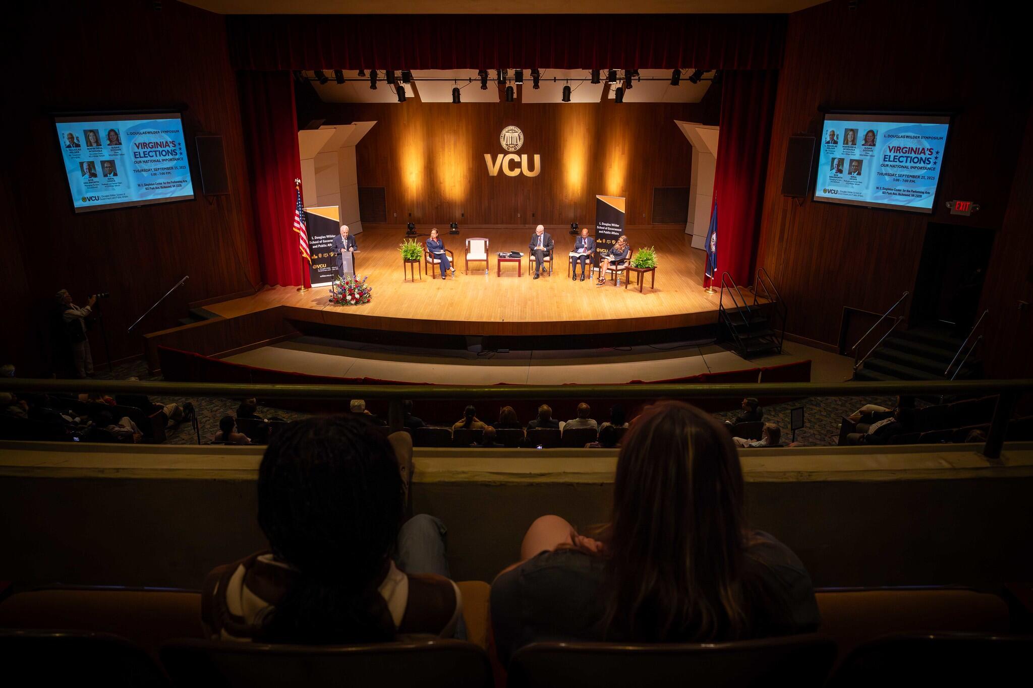 A photo of an auditorium. On the stage are four people sitting in chairs. 