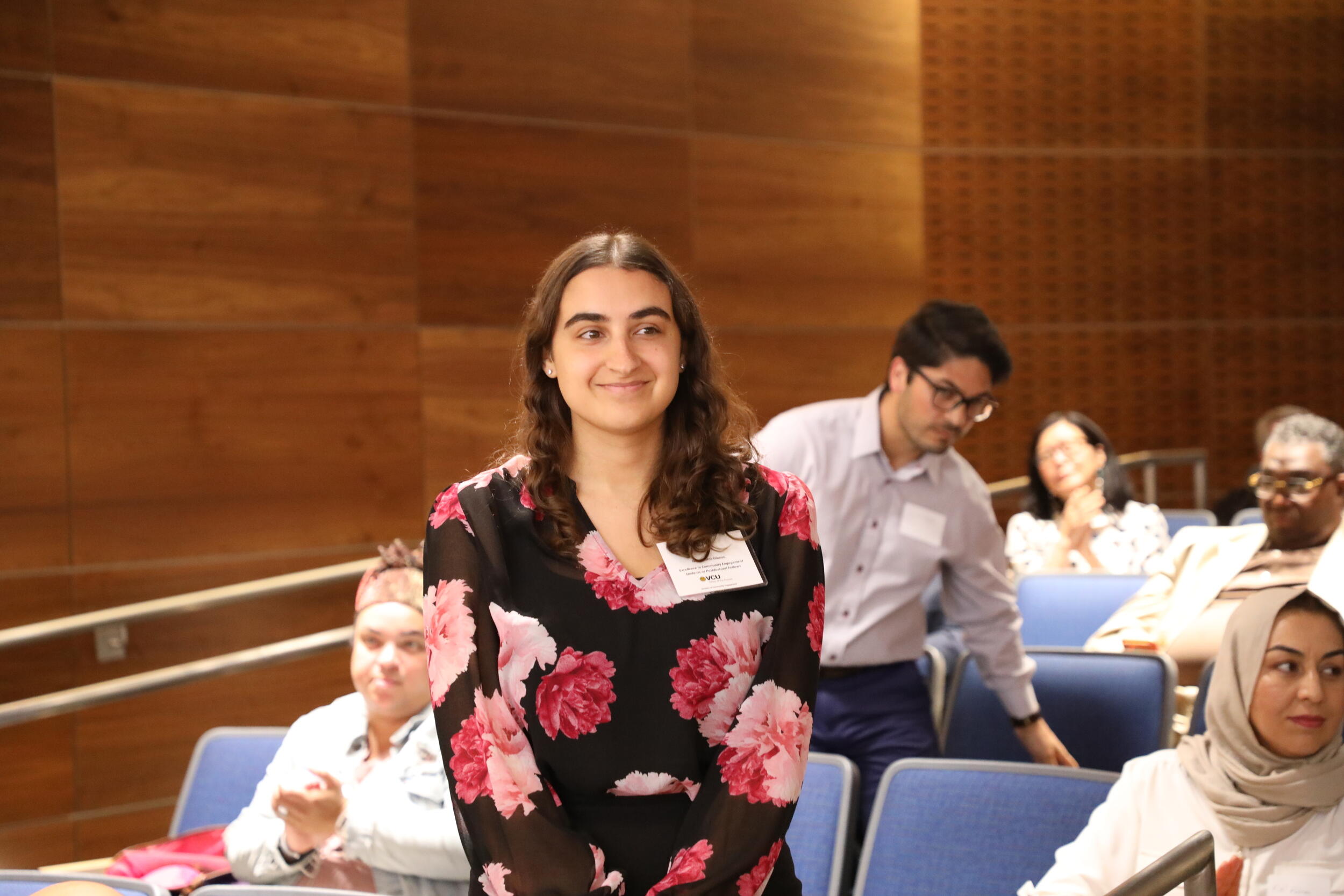 A photo of a woman from the waist up. She is standing in front of auditorium chairs. 