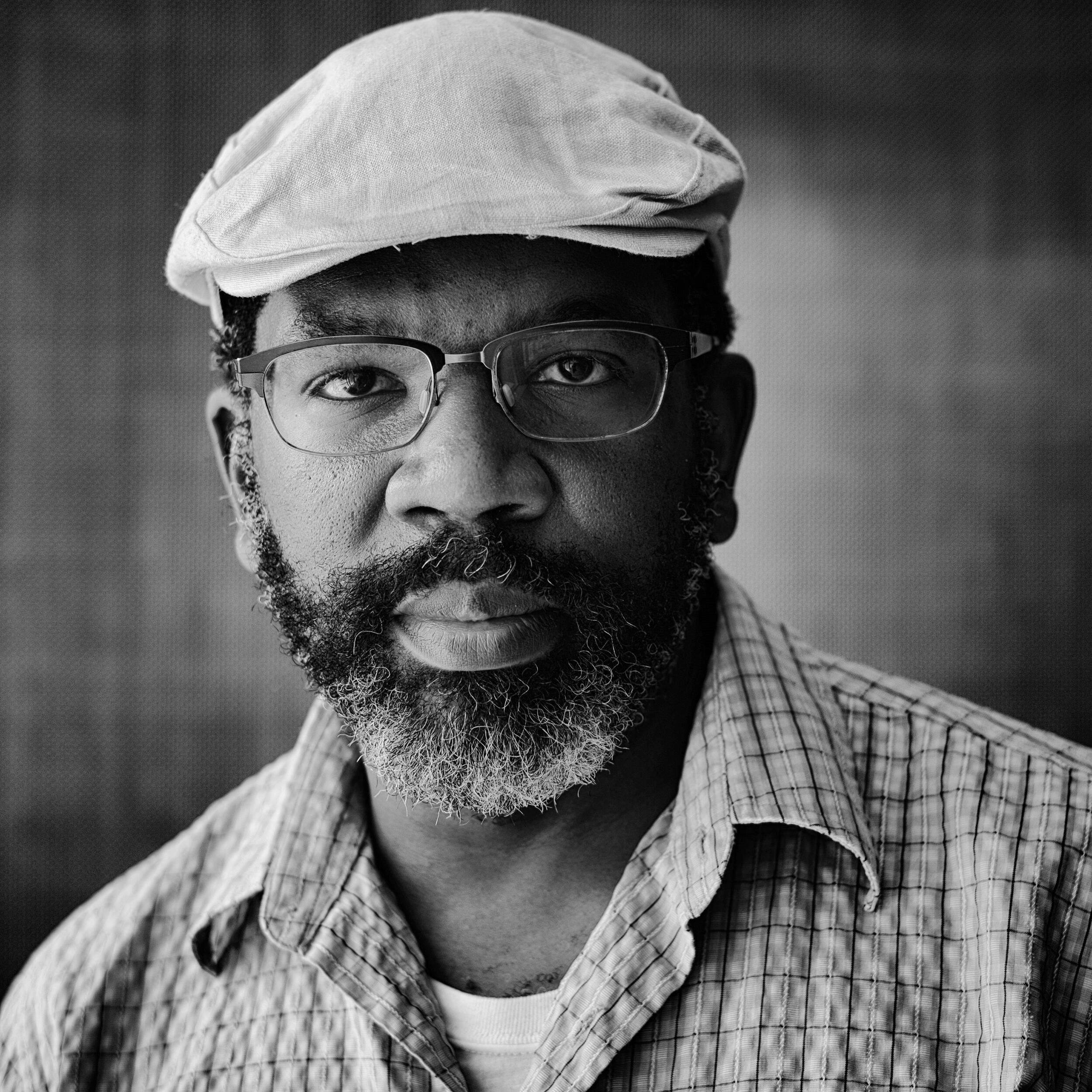 A black and white photo of a man with a short beard wearing a hat and glasses. 