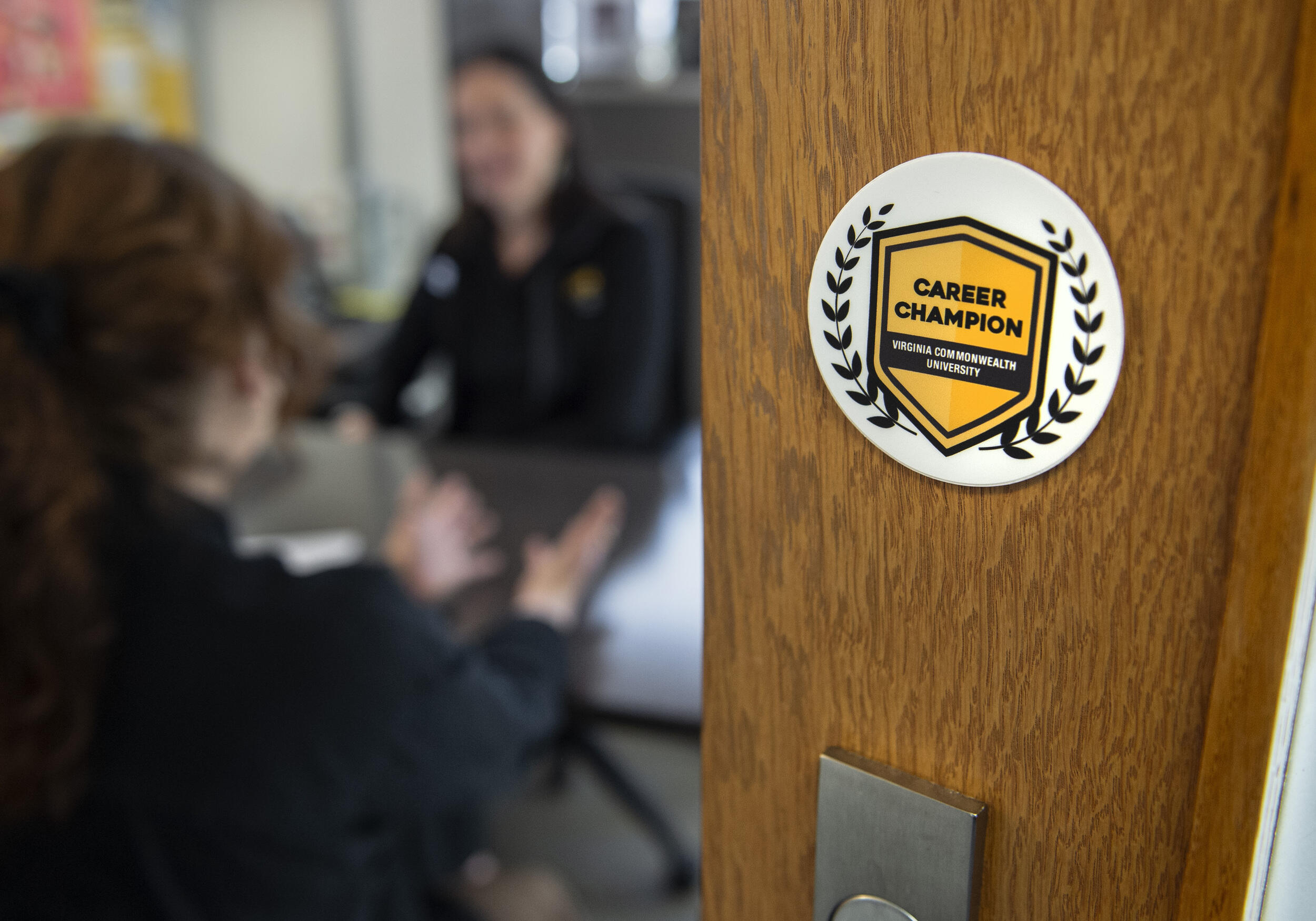 A photo of two women sitting across from each other at a desk. To the left of the women is a wooden door with a sticker that says \"CAREER CHAMPION\" in black letters on a yellow shield. 