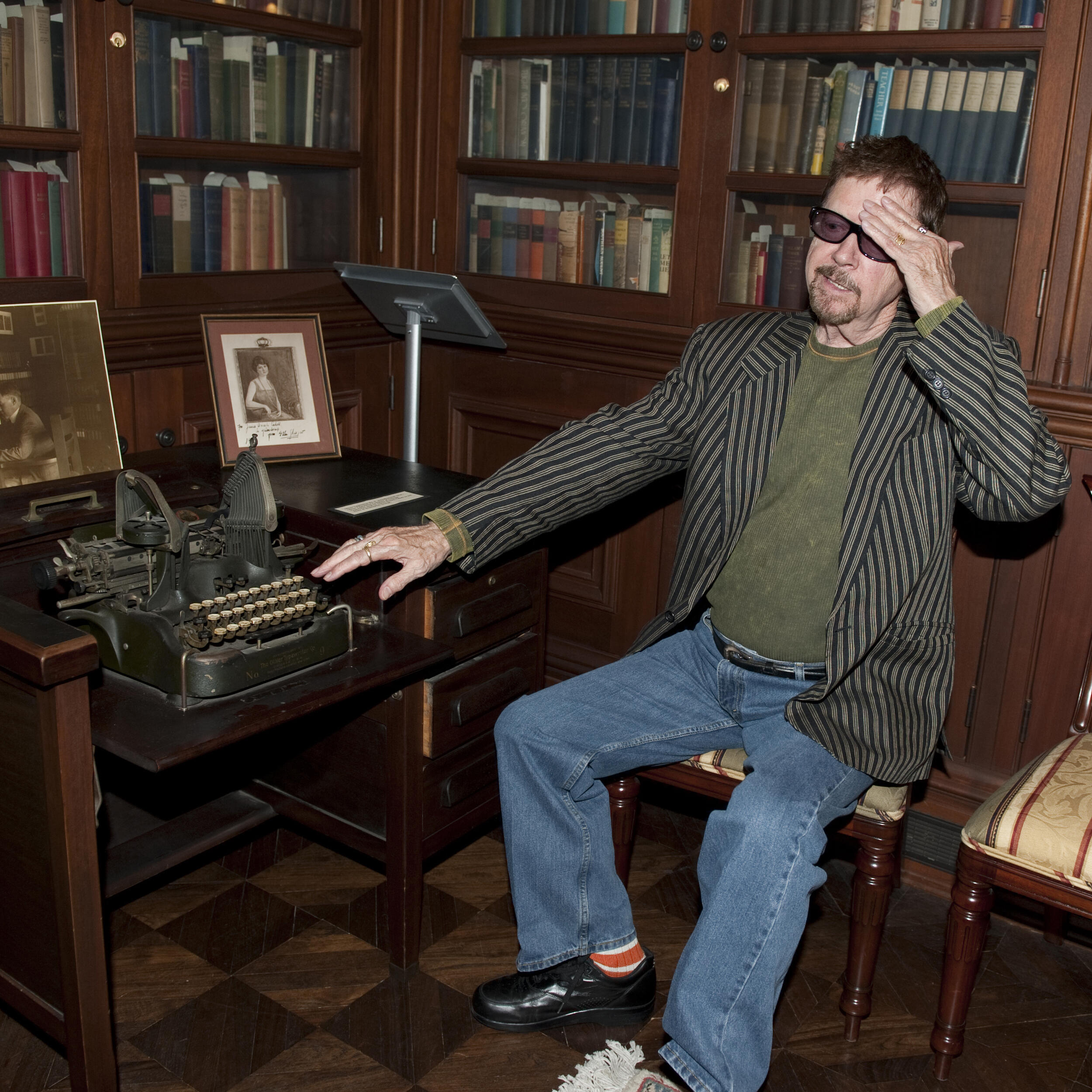 A photo of a man sitting in a chair next to a typewriter. Behind him are shelves covered in books. 