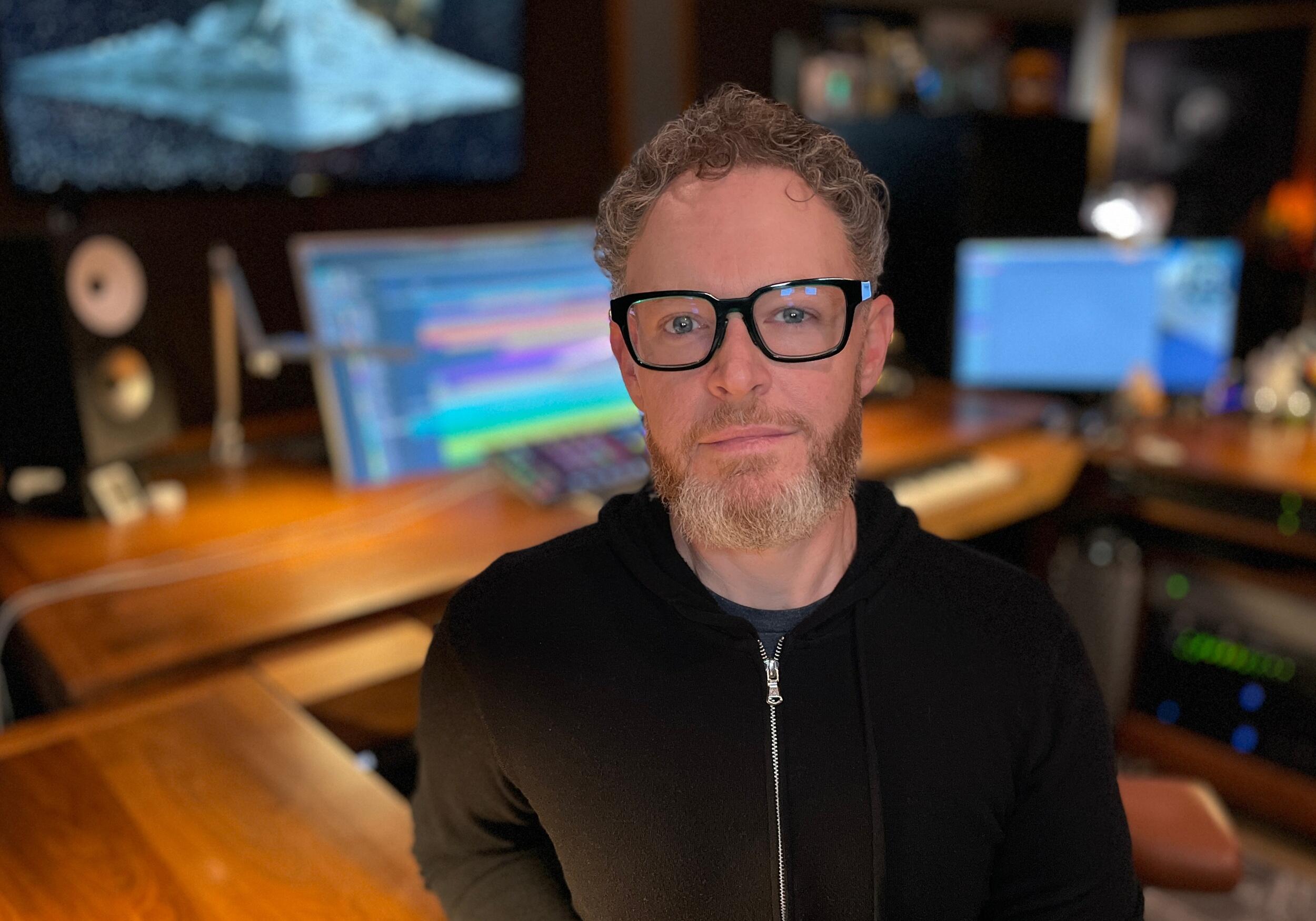 A photo of a man from the chest up standing in a music studio. 