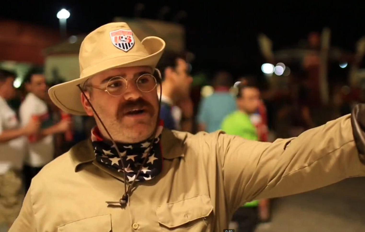 A man dressed in a hat, glasses and a mustache points.