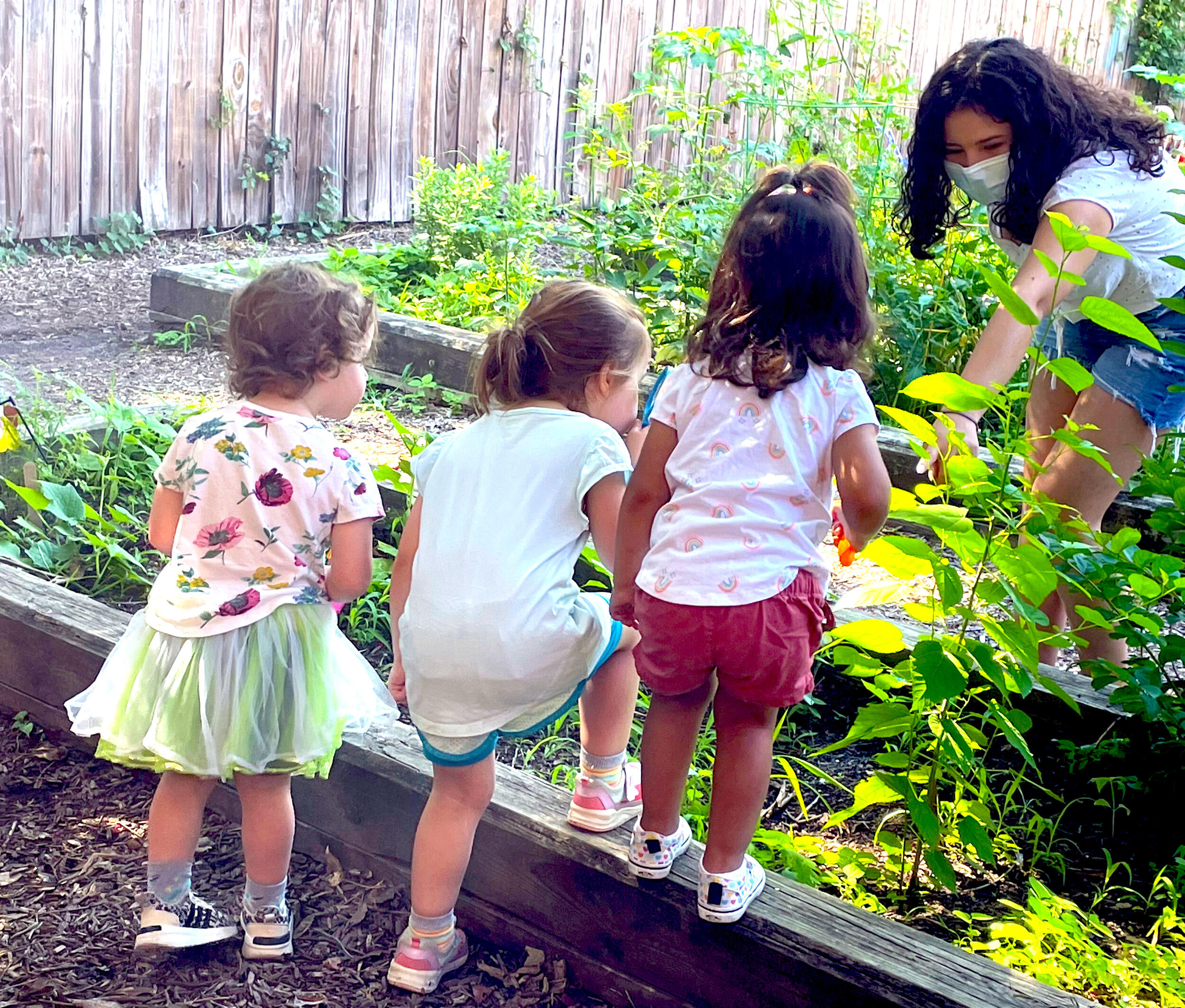 Children standing on a garden box in front of a woman wearing a face mask 