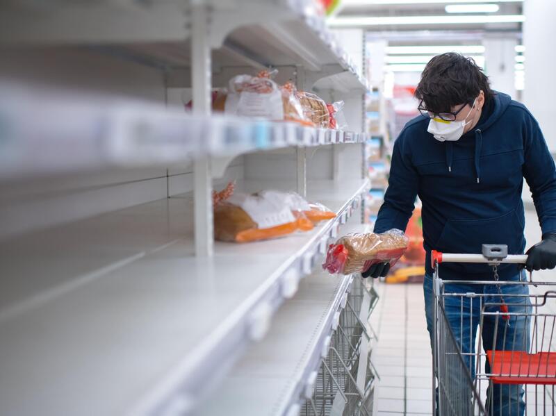 Shopper in a face mask picks up a loaf of bread from mostly empty shelves.