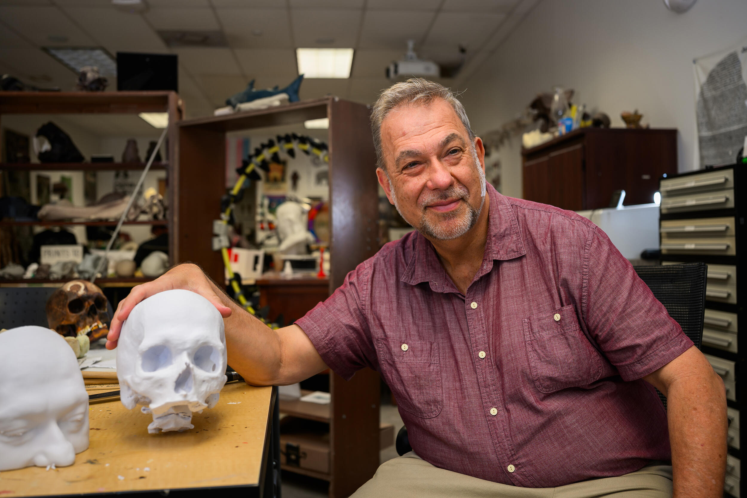 A photo of a man sitting next to a table with a 3D prited human skull and upper half of a human's head on it. 