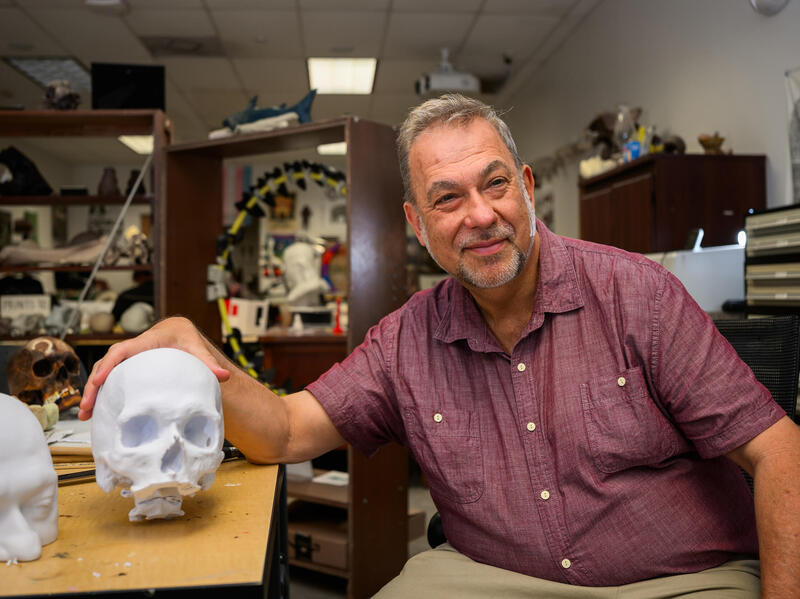 A photo of a man sitting next to a table with a 3D prited human skull and upper half of a human's head on it. 