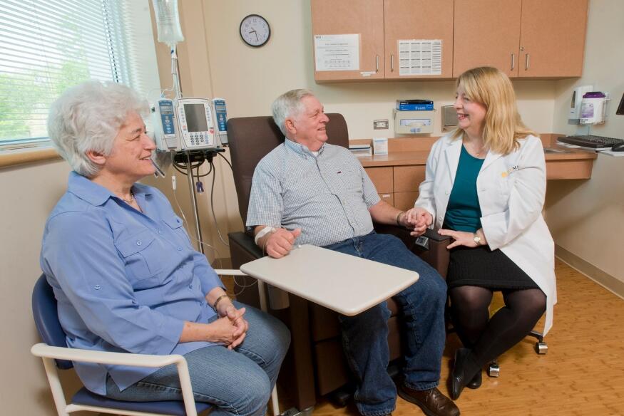 A photo of three people sitting in a medical examination room. On the far right is a doctor sitting on a rolling stool. In the center is a man sitting on an examination chair. To the far left is a woman sitting in a chair. 