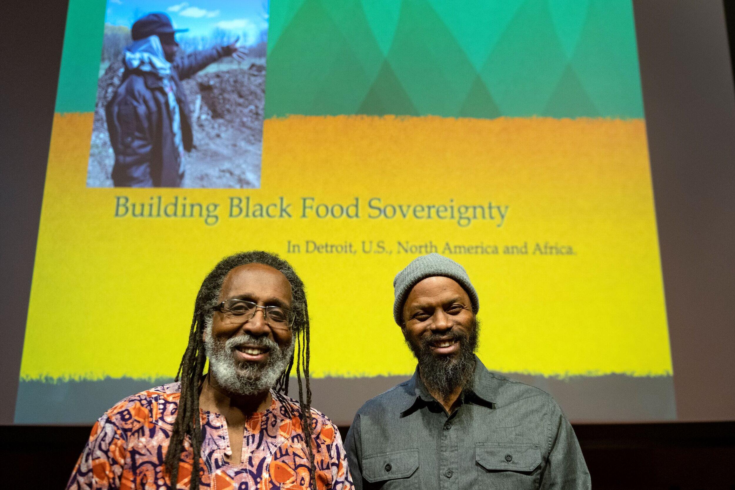 Two men standing in front of a screen that says \"Building Black Food Sovereignty in Detroit, U.S., North America and Africa\"