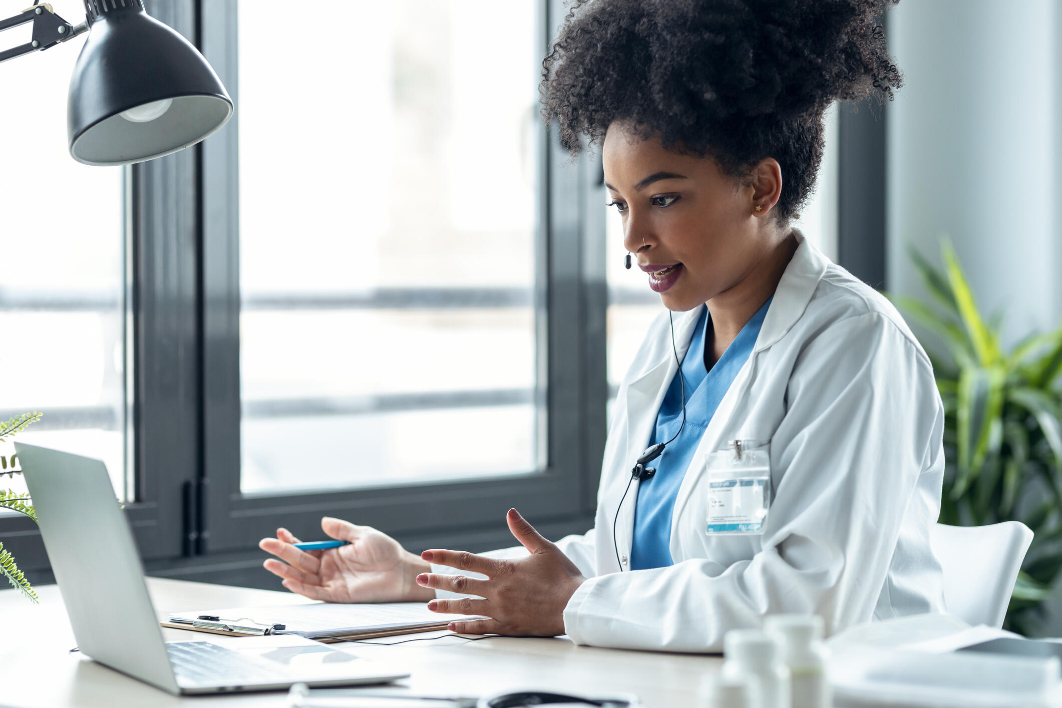 A photo of a doctor sitting at a desk with a laptop in front of her. She is speaking into a headset and gesturing her hands in front of her.