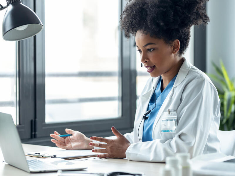 A photo of a doctor sitting at a desk with a laptop in front of her. She is speaking into a headset and gesturing her hands in front of her. 