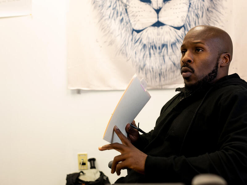 A photo of a man from the waist up. He is talking and holding a stack of paper. 