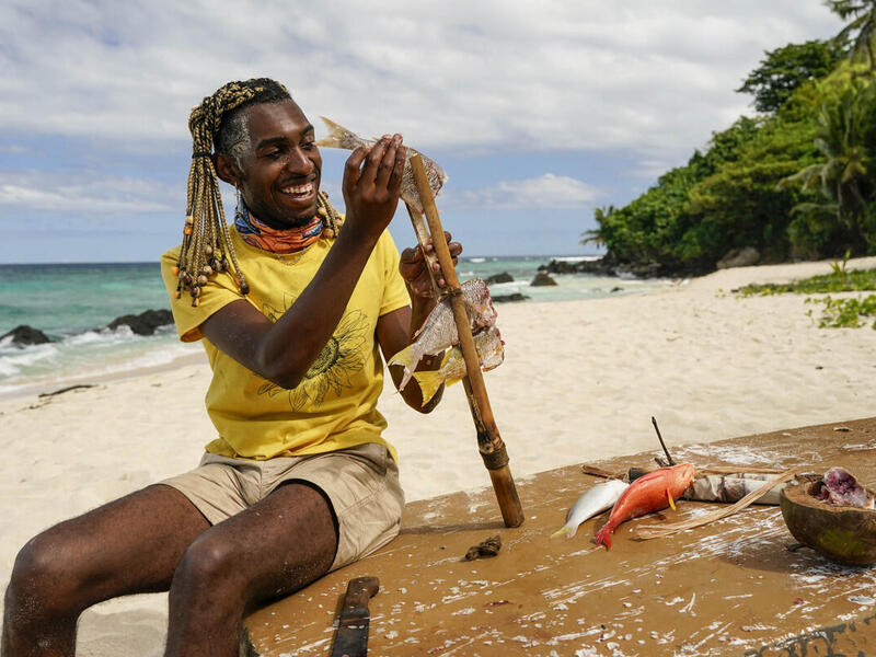 A photo of a man sitting on a rock on a beach. He is holding a stick with a fish on it. 