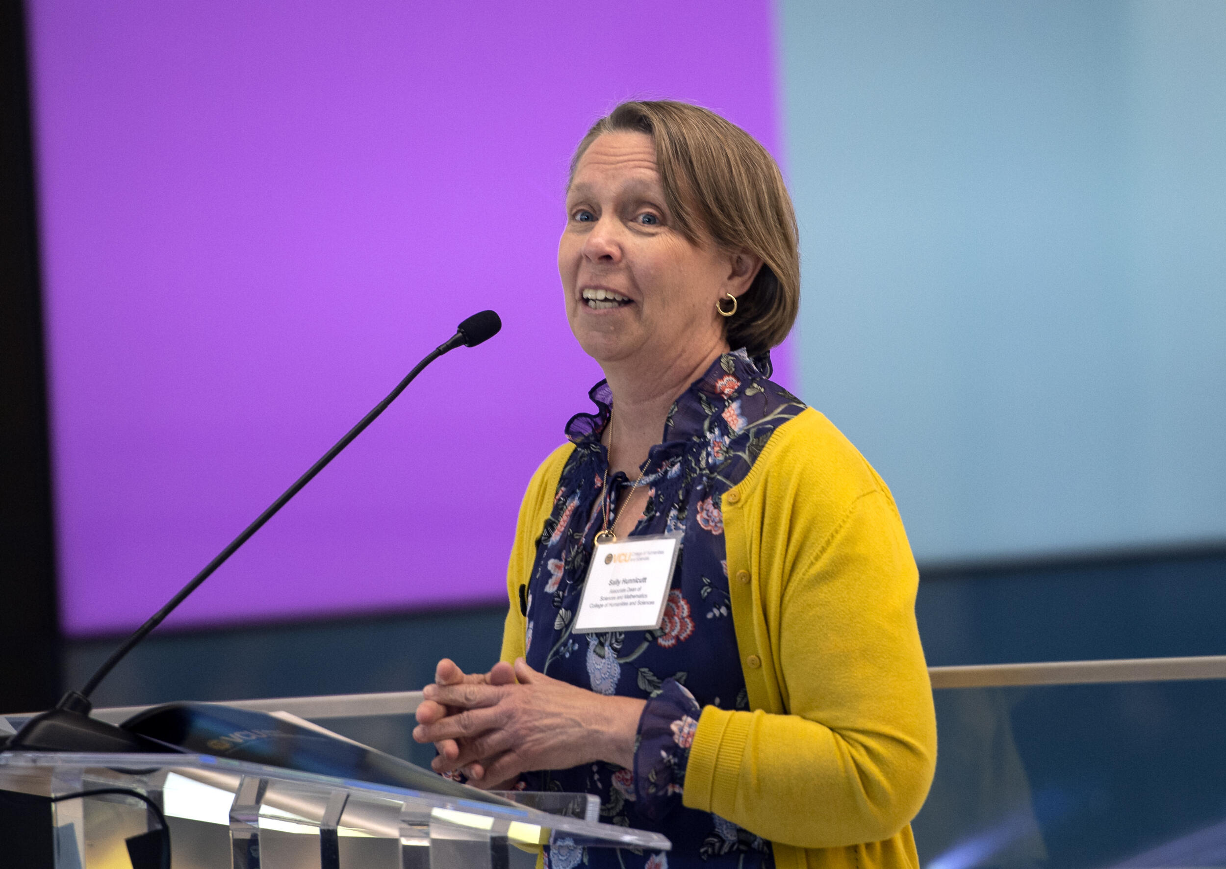 A photo of a woman from the waist up. She is standing behind a podium and speaking into a microphone