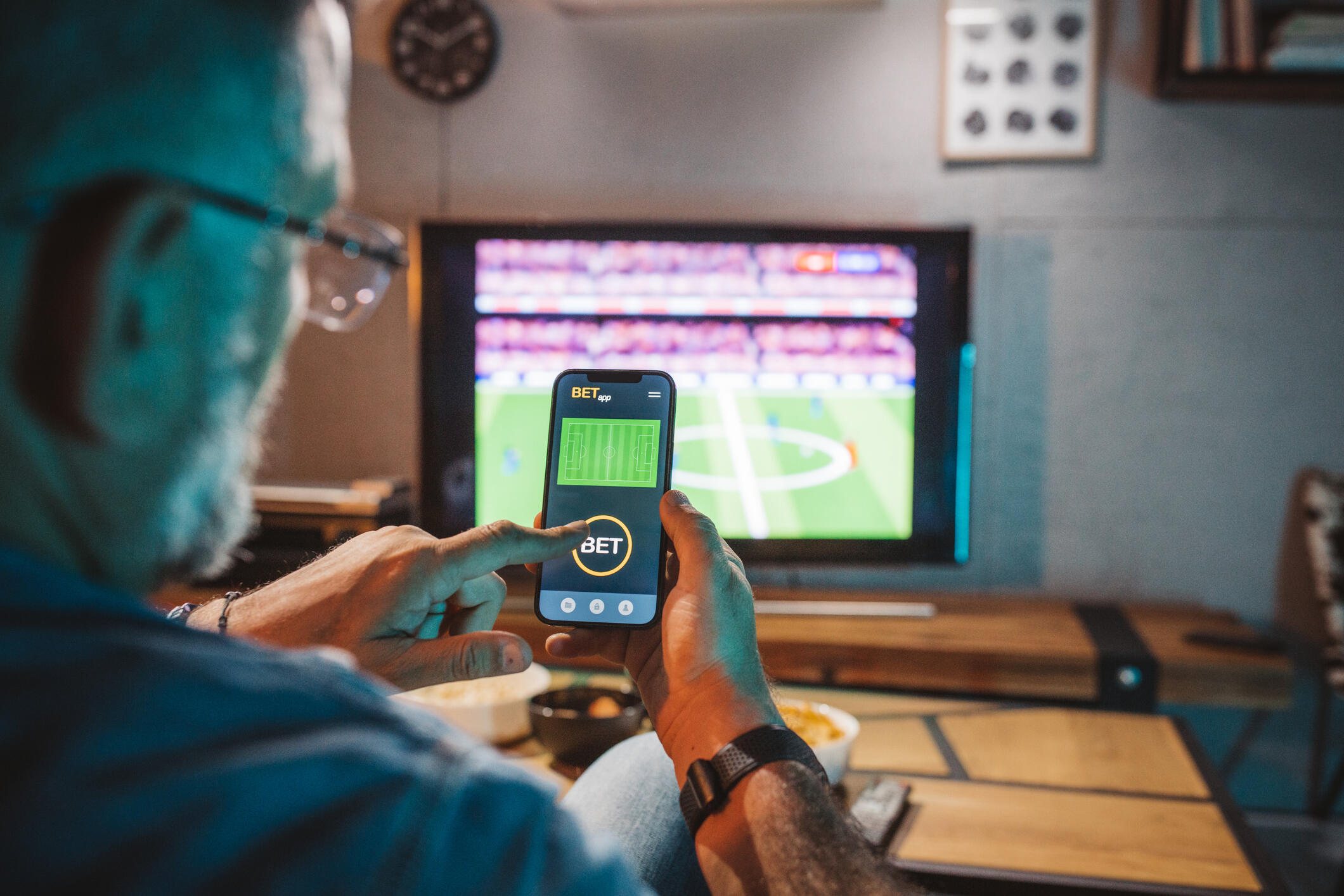 A photo of a man looking at a sports betting app on his smart phone while a football game is playing on the TV in front of him. 