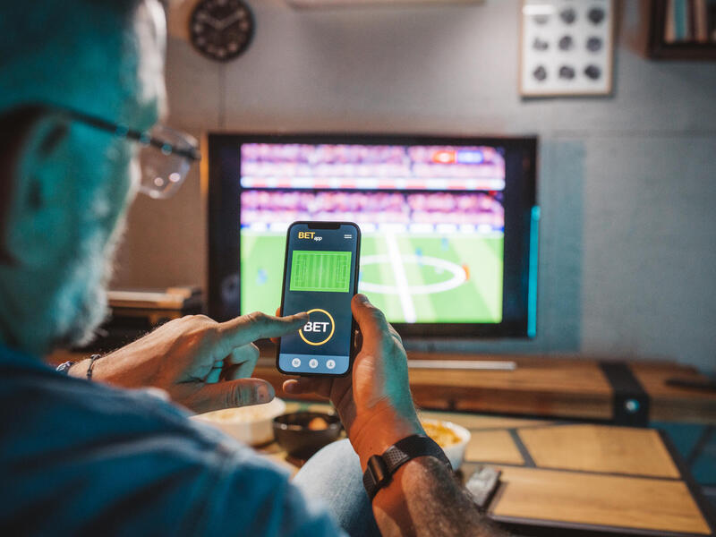 A photo of a man looking at a sports betting app on his smart phone while a football game is playing on the TV in front of him. 