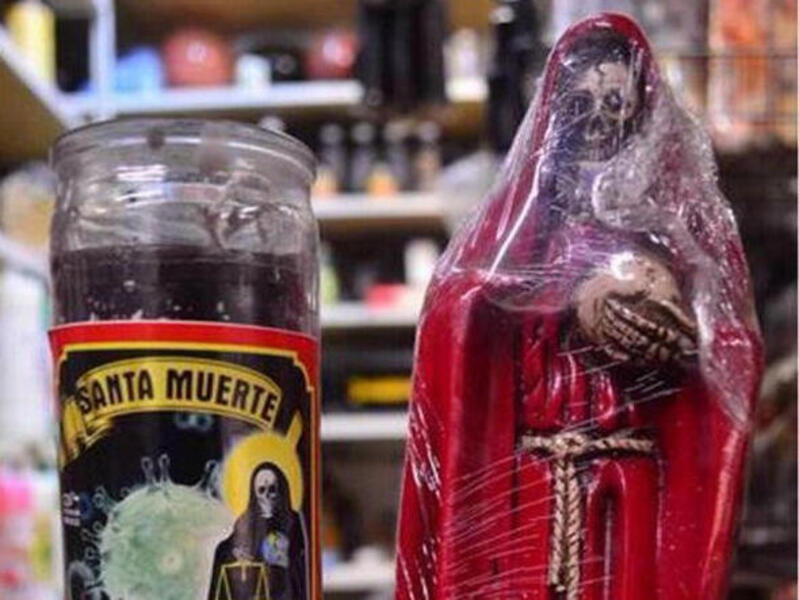 Santa Muerte's visage, typically depicted in symbolic votive candles (red for love, gold for prosperity, purple for healing), has been in such high demand as a symbol of healing during the pandemic that sellers in religious goods stores in Mexico have created candles like this one (left, next to a small statue of Santa Muerte) in a new color — a deep shade of mauve — specifically for coronavirus healing.