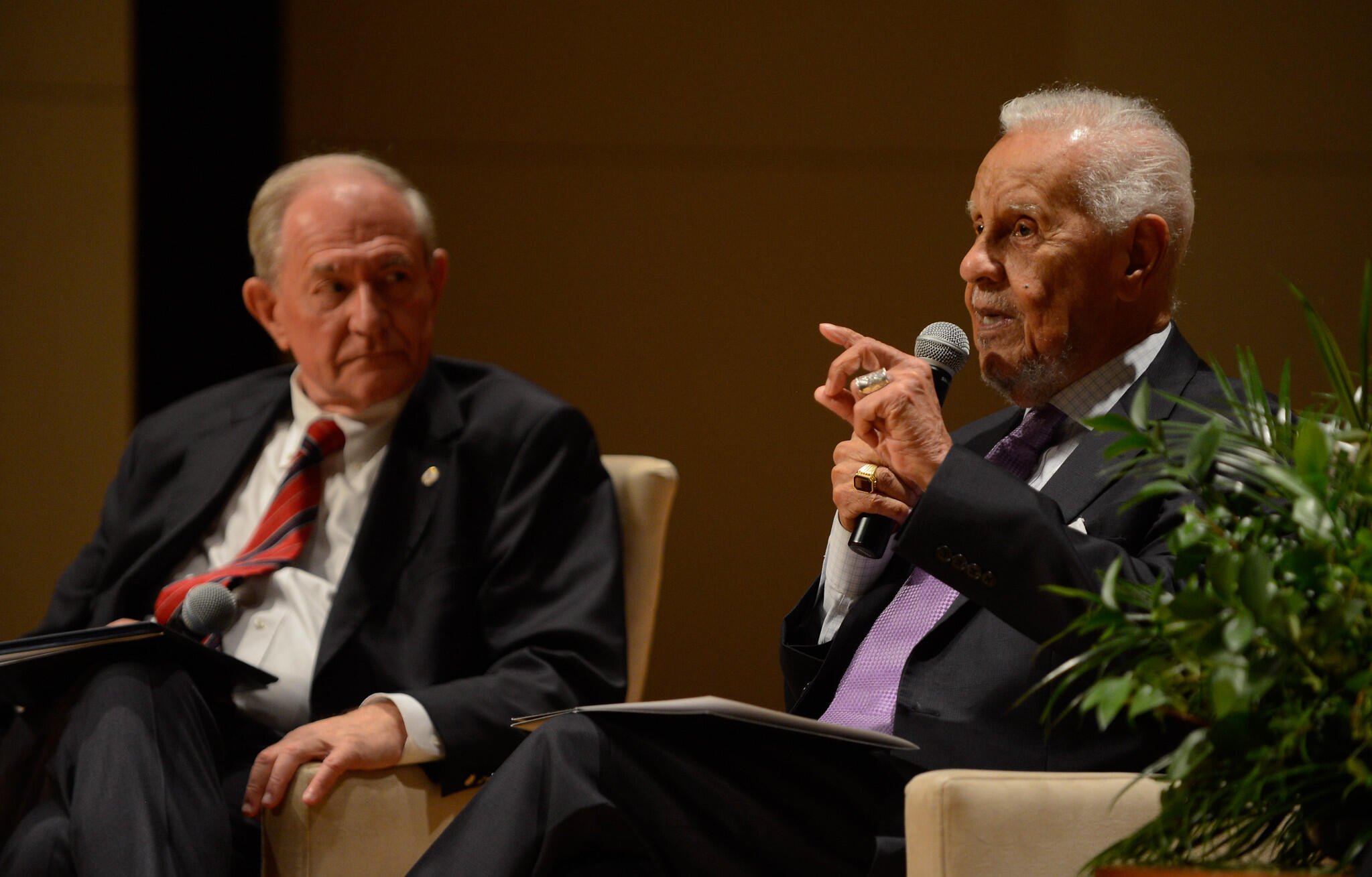 A photo of two men sitting in arm chairs. The man on the right is speaking into a microphone. 