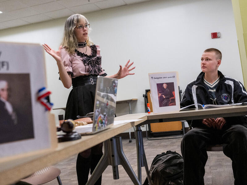 A person stands and speaks, gesturing with their hands, while another person sits nearby at a table and watches.