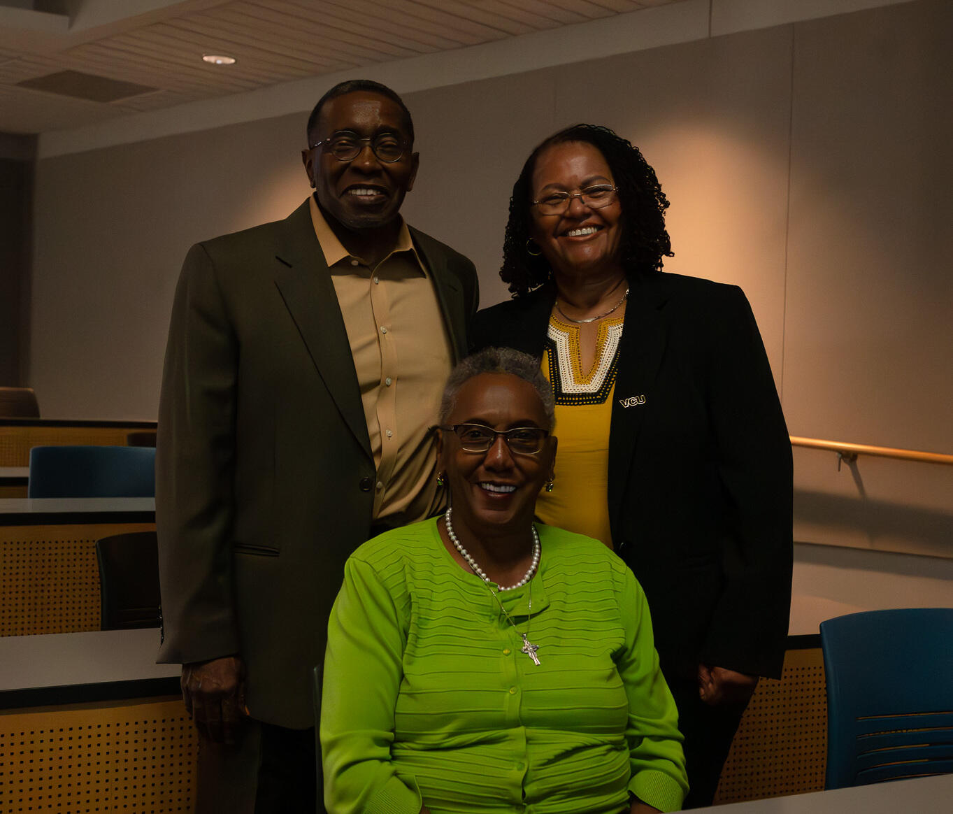 One woman sits at a table in a green jacket and a man and a woman stand behind her.