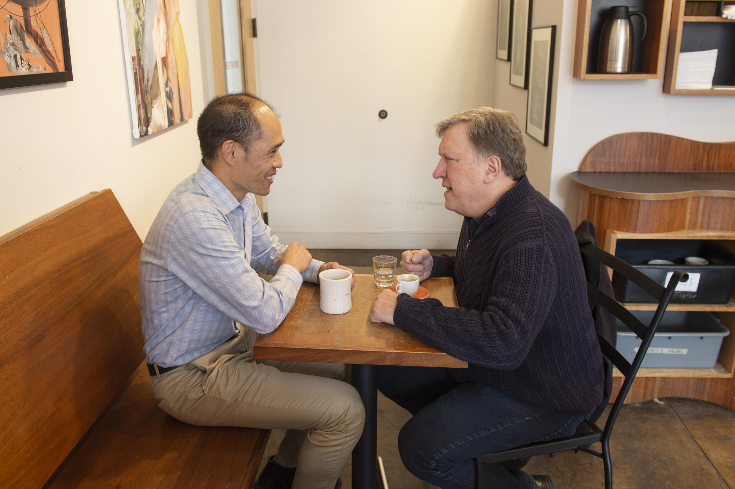 Two men sitting across from each other at a table with a coffee and expresso cup in front of them. 