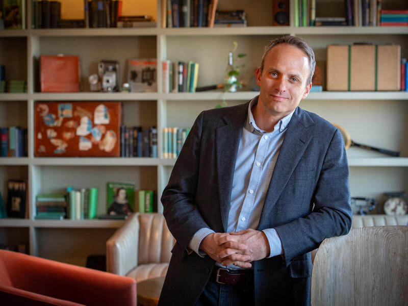 A photo of a man from the waist up standing in front of a book shelf. 