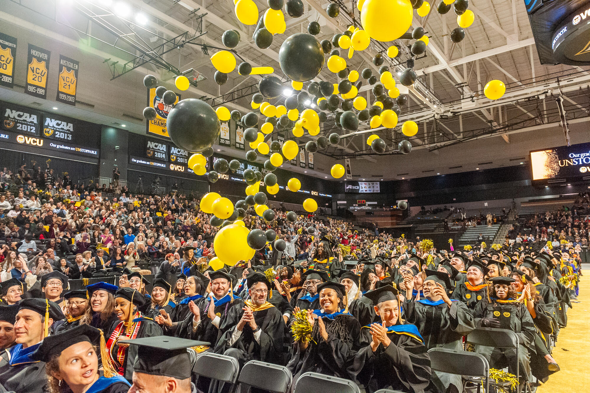 A photo of black and yellow balloons falling down on a group of students wearing graduation cap and gowns. The students are sitting in rows of chairs. Around the students are bleachers filled with people sitting in them.