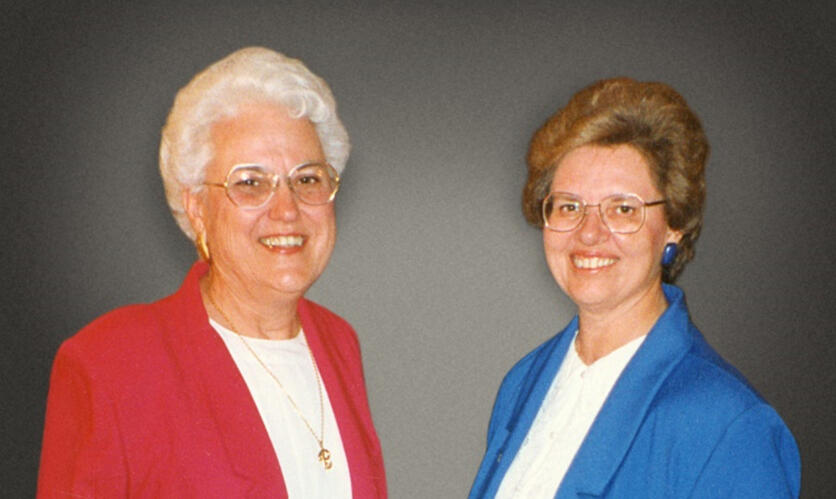 Two women, one in a red blazer at left and one in a blue blazer at right.