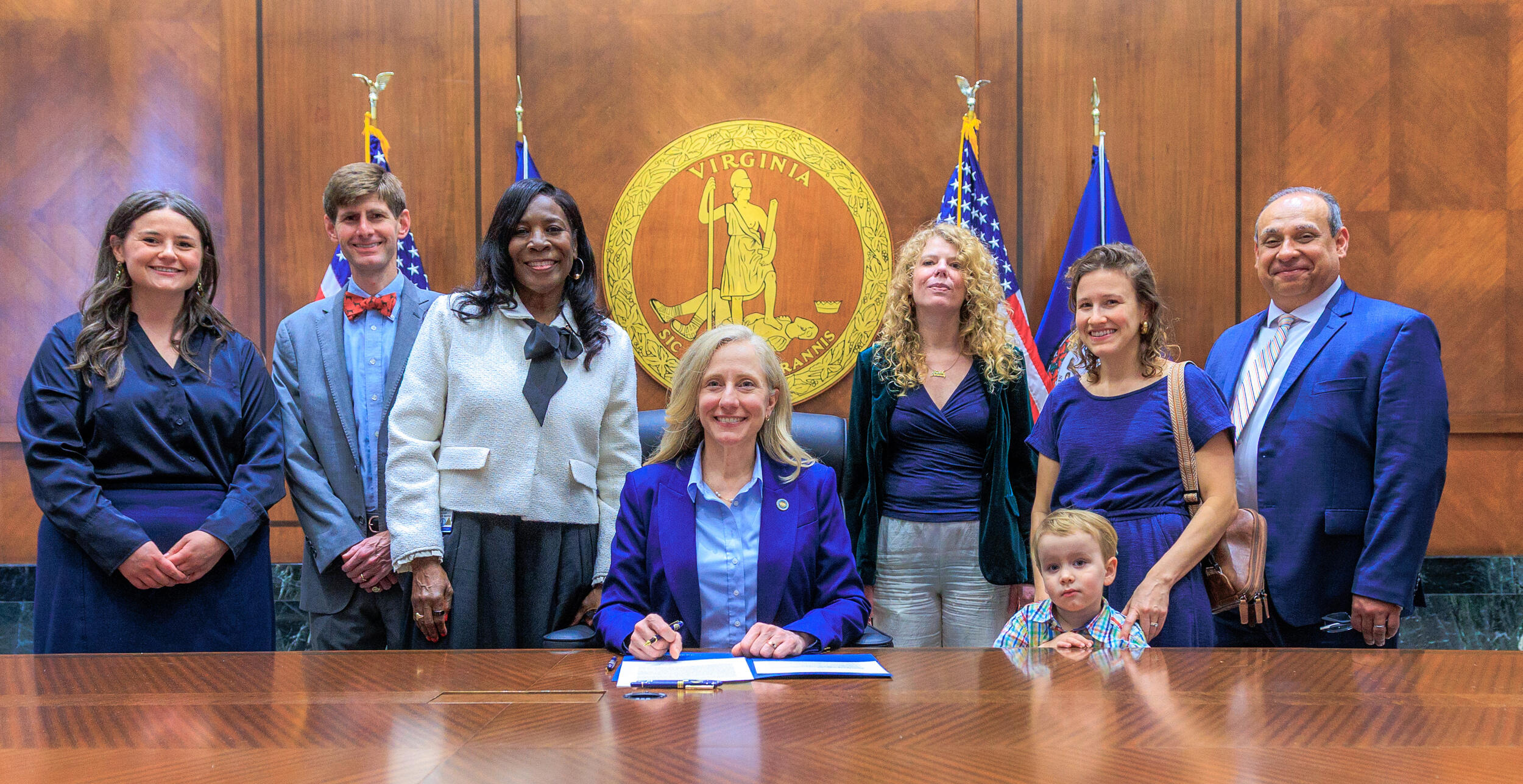 A group photo of three adults standing on either side of a woman sitting at a table. On the right side, there is is also a child lenaing against the table the woman is sitting at. 