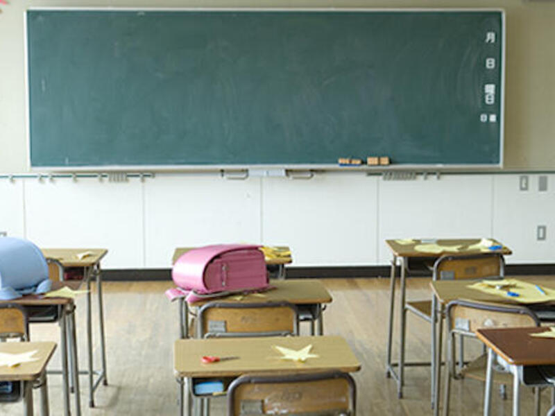 A classroom with wooden desks facing a green chalkboard. Two of the desks have backpacks on them. 