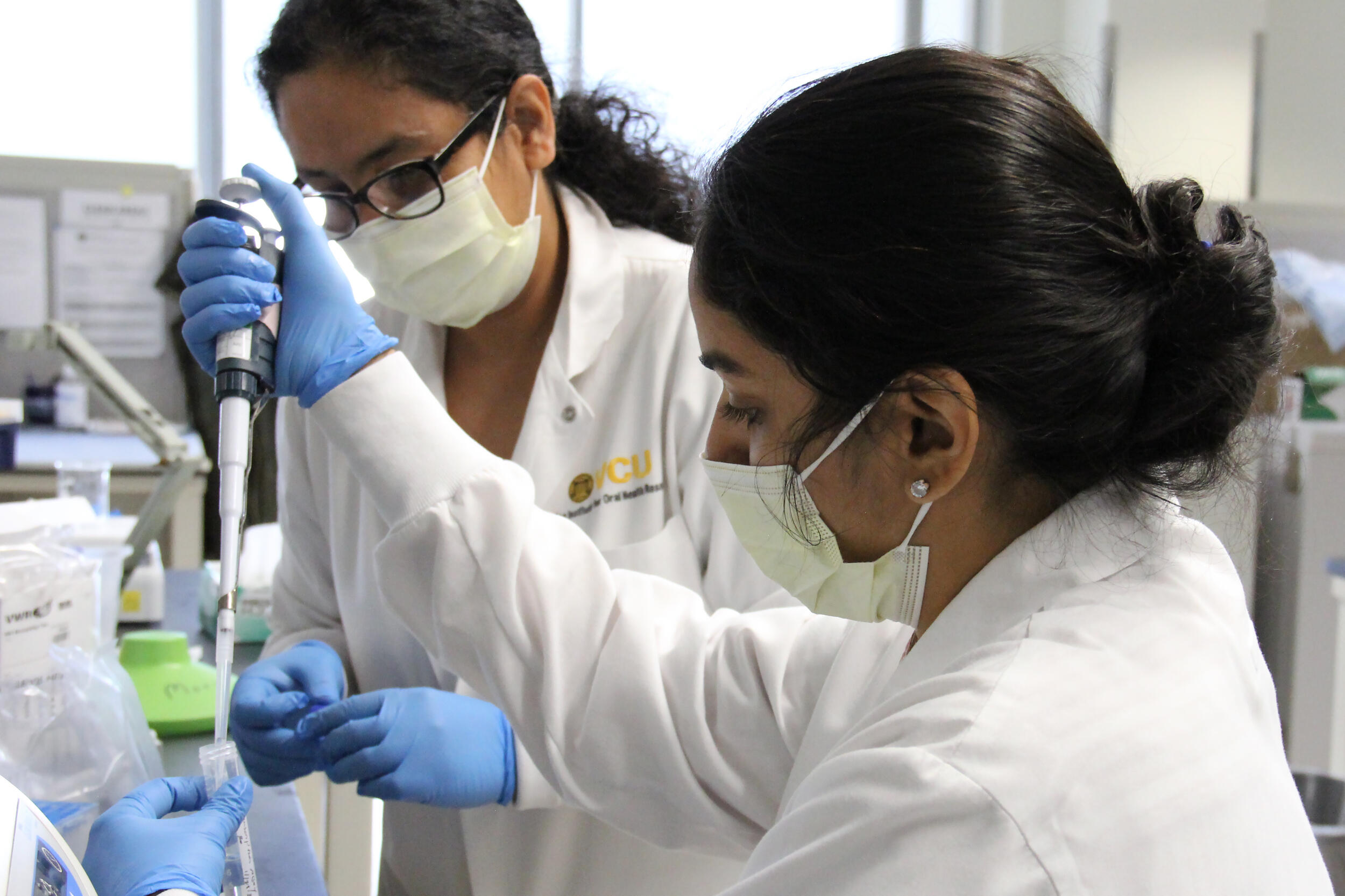 Two women wearing lab coats, face masks, and rubber gloves in a research lab. 