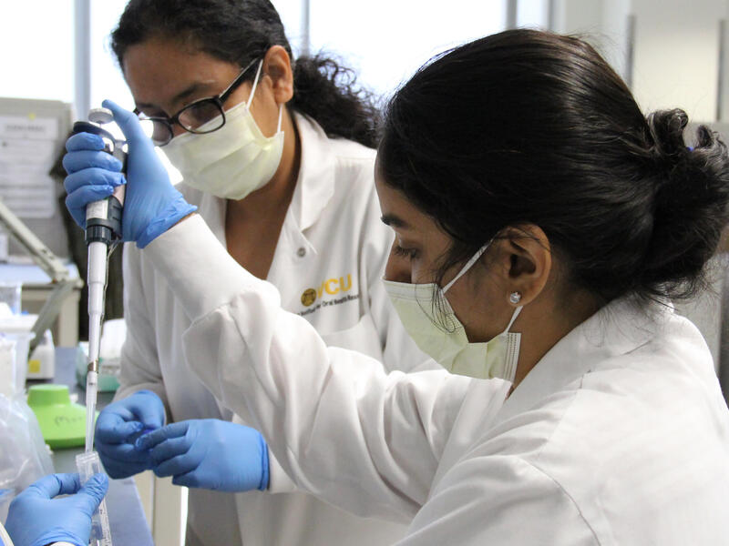 Two women wearing lab coats, face masks, and rubber gloves in a research lab. 