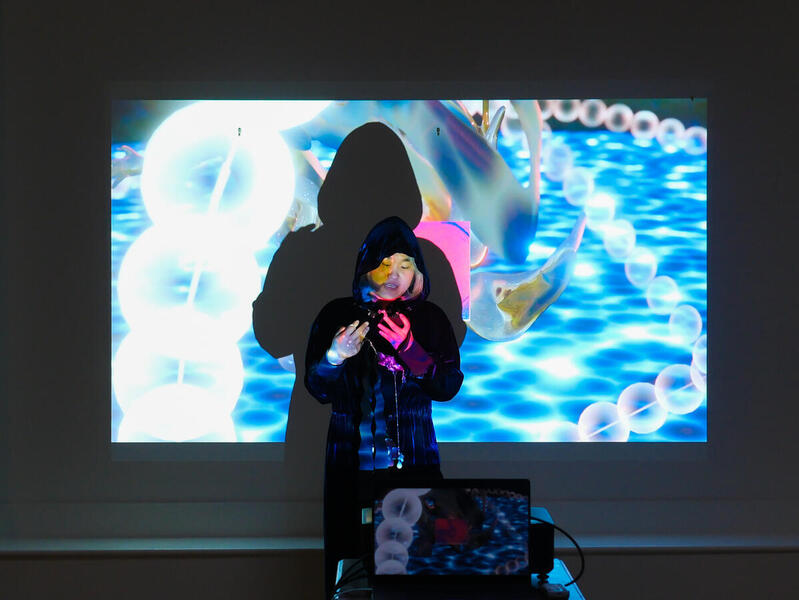 A woman standing in front of a projector that is projecting an image of a 3D fish in an abstract environment.