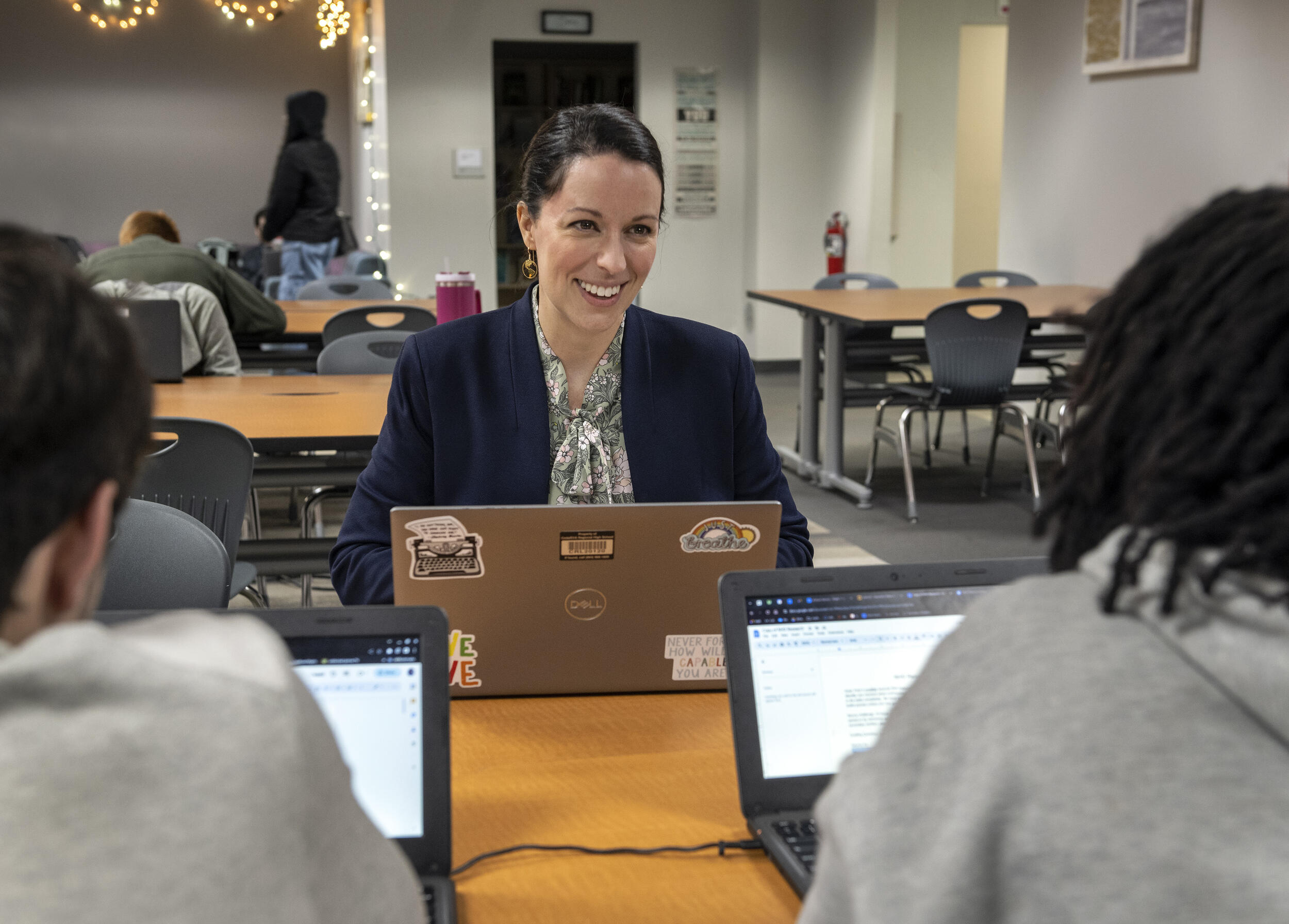 A woman sitting with a laptop smiling at two students who are also on their laptops. 